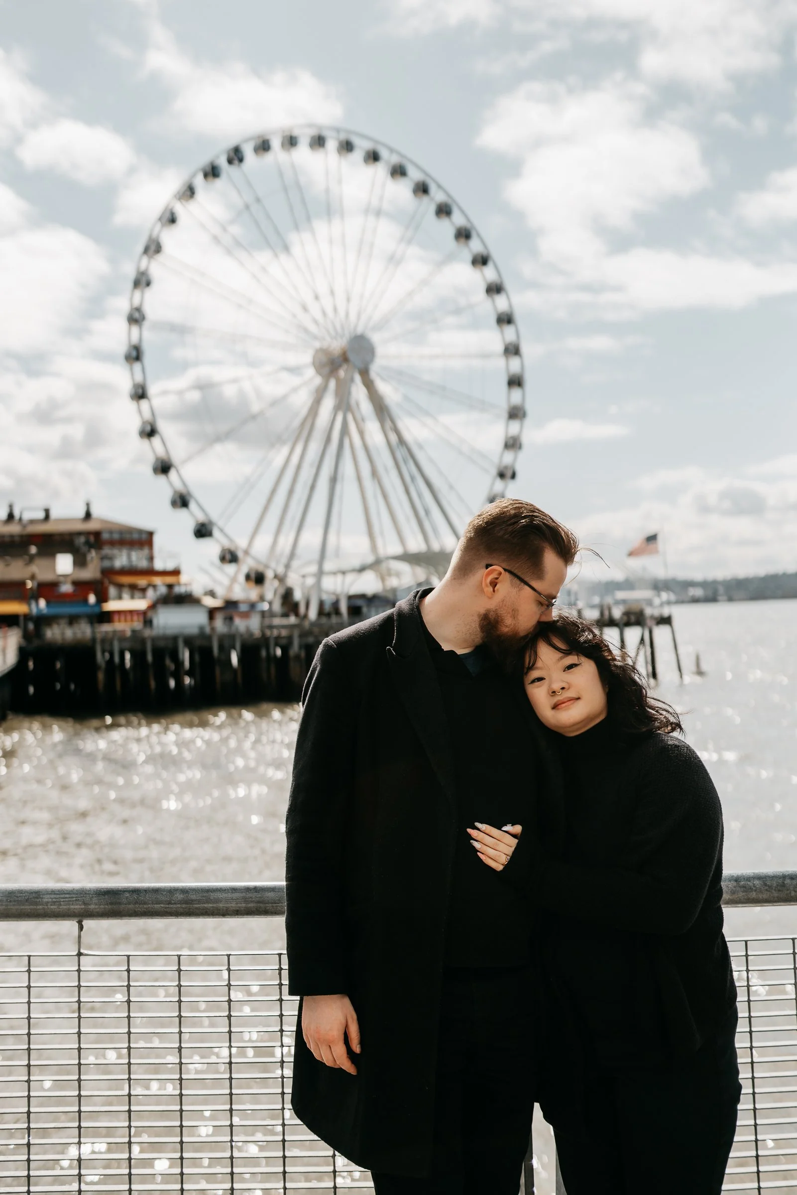 A couple embracing near a waterfront with a Ferris wheel in the background. The man is kissing the woman on the forehead, and both are wearing dark clothing. The scene is outdoors on a partly cloudy day.