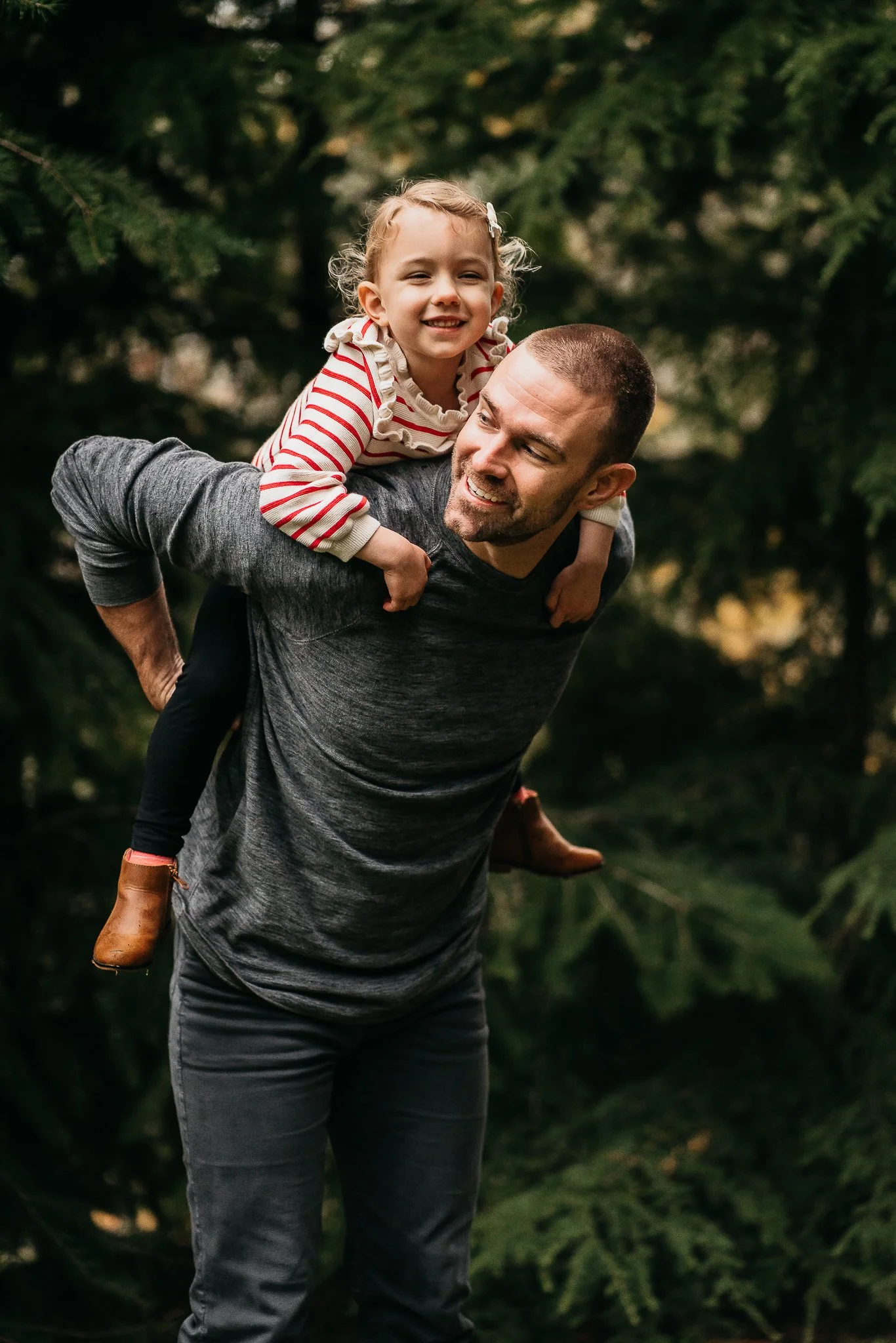 A man giving a piggyback ride to a young girl in a forest setting, both smiling and enjoying the moment.
