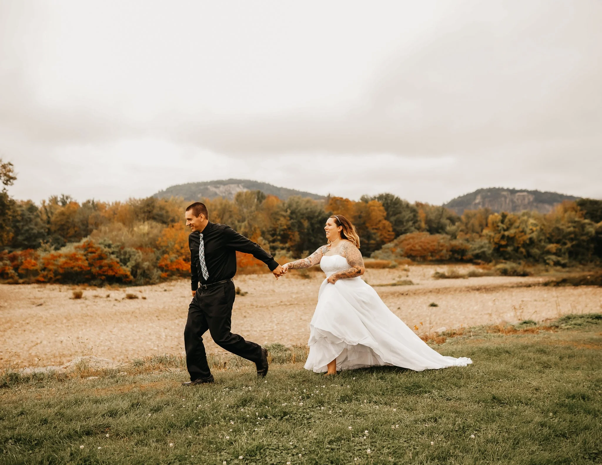 A groom and bride holding hands and running outdoors on a cloudy day, with the bride in a white wedding dress and the groom in a black shirt and tie, in a scenic landscape with autumn-colored trees and hills in the background.