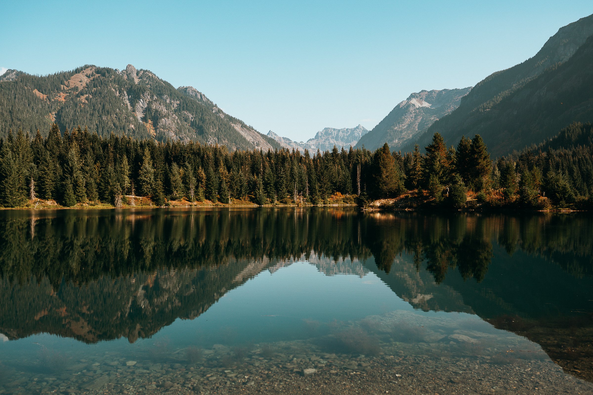 A calm mountain lake reflecting tall pine trees and rugged mountains under a clear blue sky.
