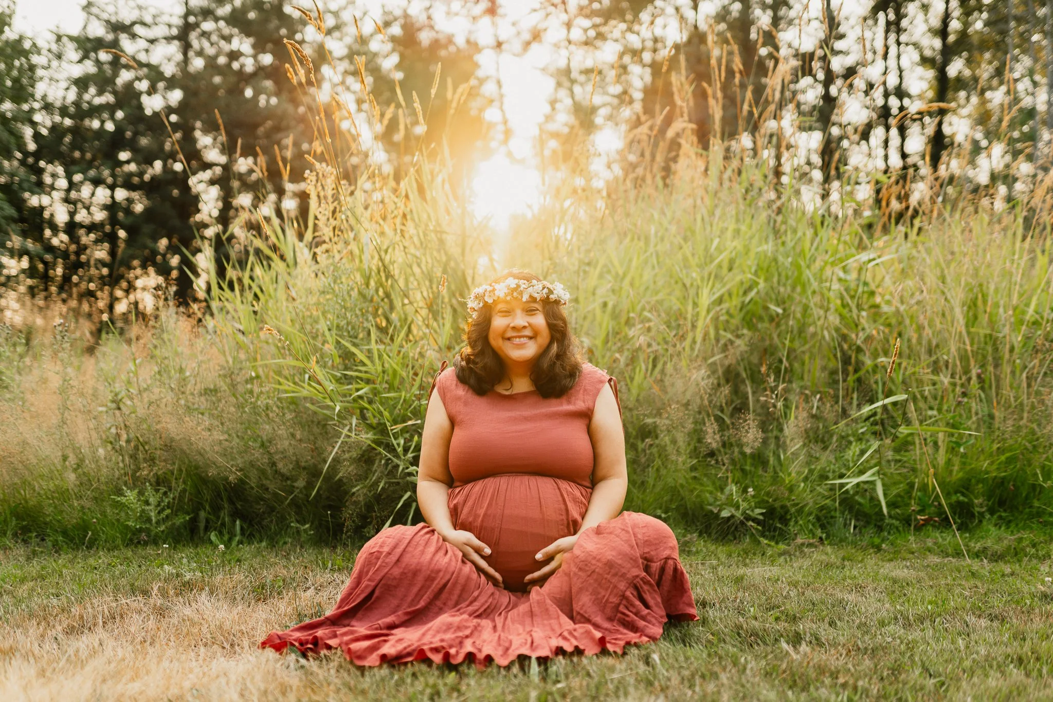Pregnant woman sitting outdoors on grass, wearing a flowing rust-colored dress and a floral crown, smiling with her hands resting on her belly, surrounded by tall grass and trees in the background during sunset.