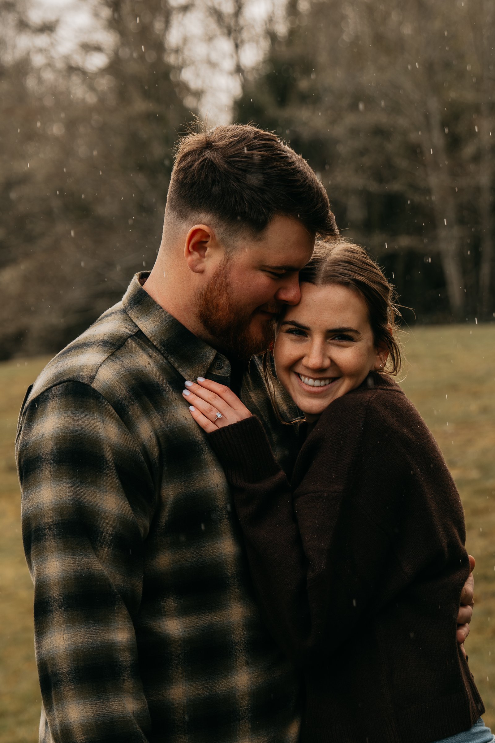 A smiling woman and a man with a beard embracing outdoors in a park, with trees and falling rain in the background.