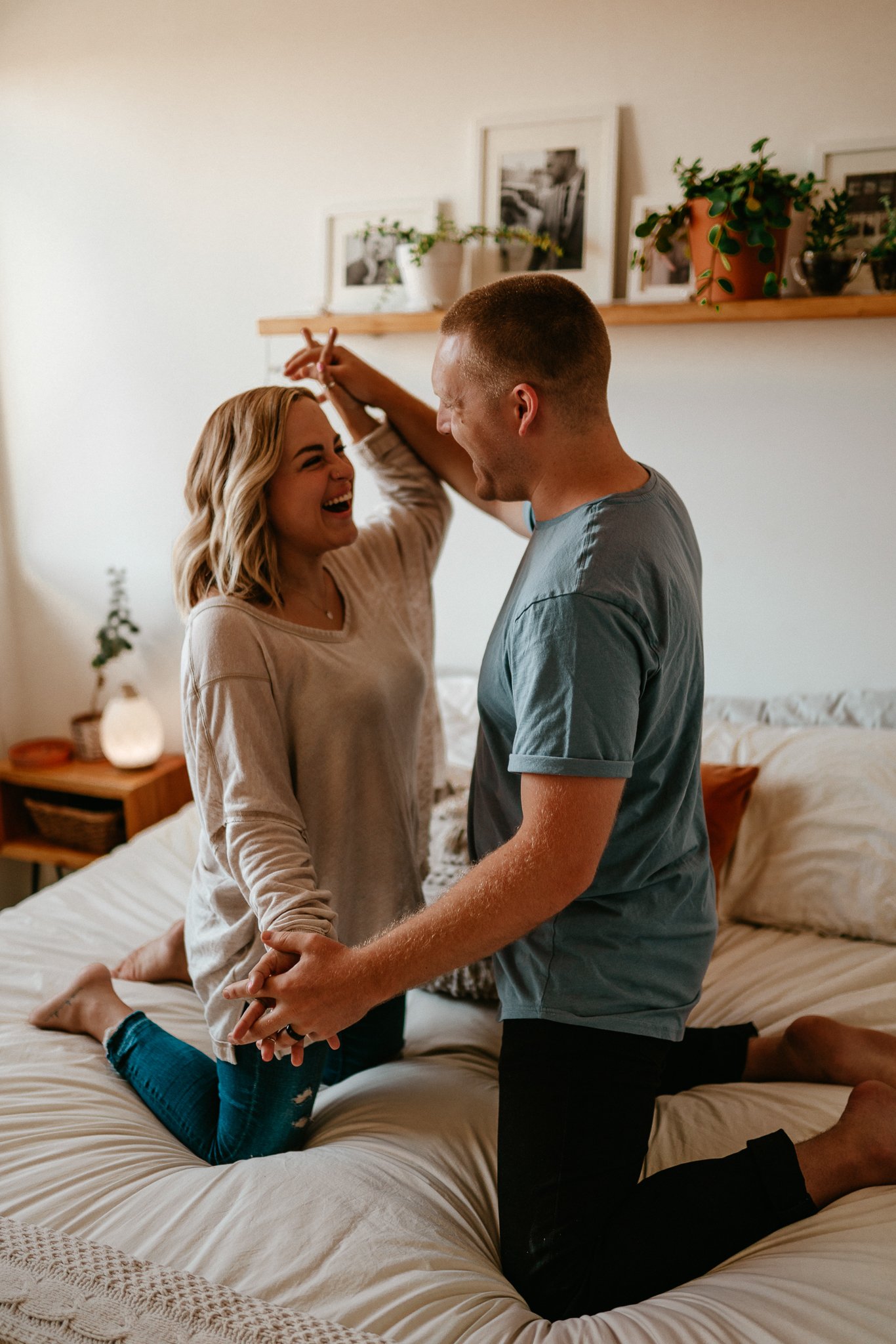 A couple is on a bed, laughing and holding hands, with one person on their knees and the other sitting on the bed, in a cozy, decorated bedroom.