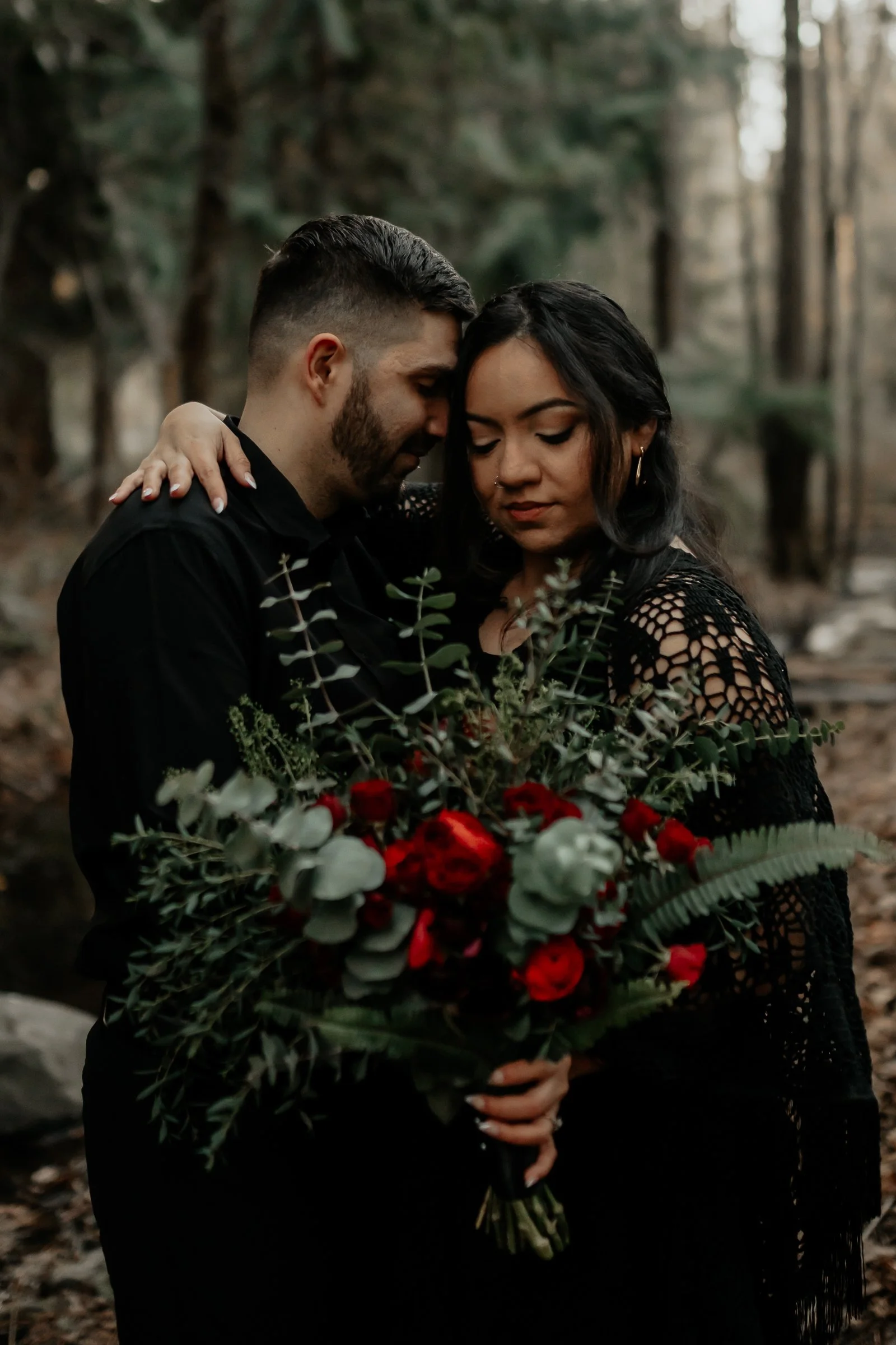 A couple embraces in a forest, holding a large bouquet of red and white flowers with green foliage.