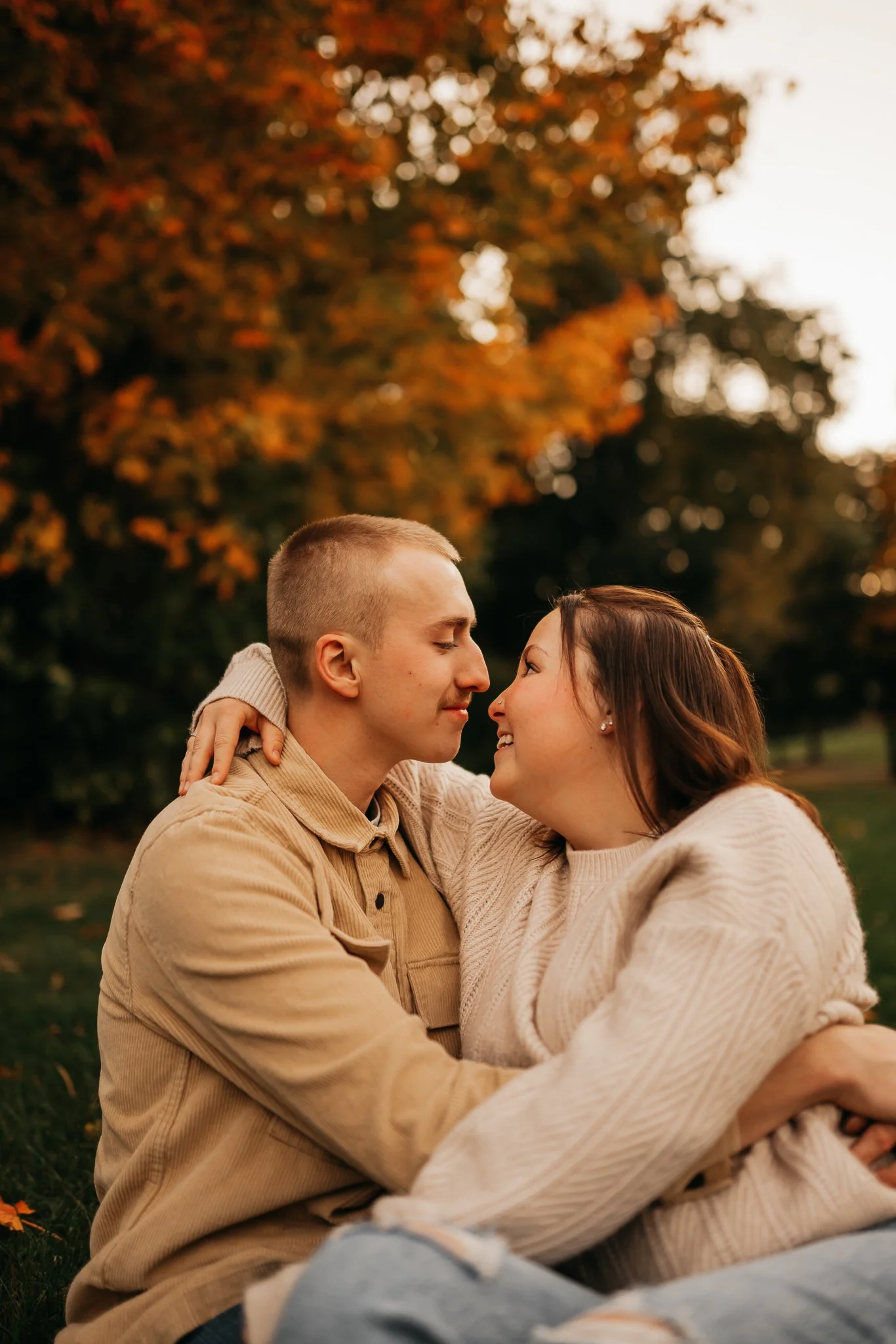 A couple sits close together outdoors during autumn, smiling and about to kiss, with fall foliage in the background.
