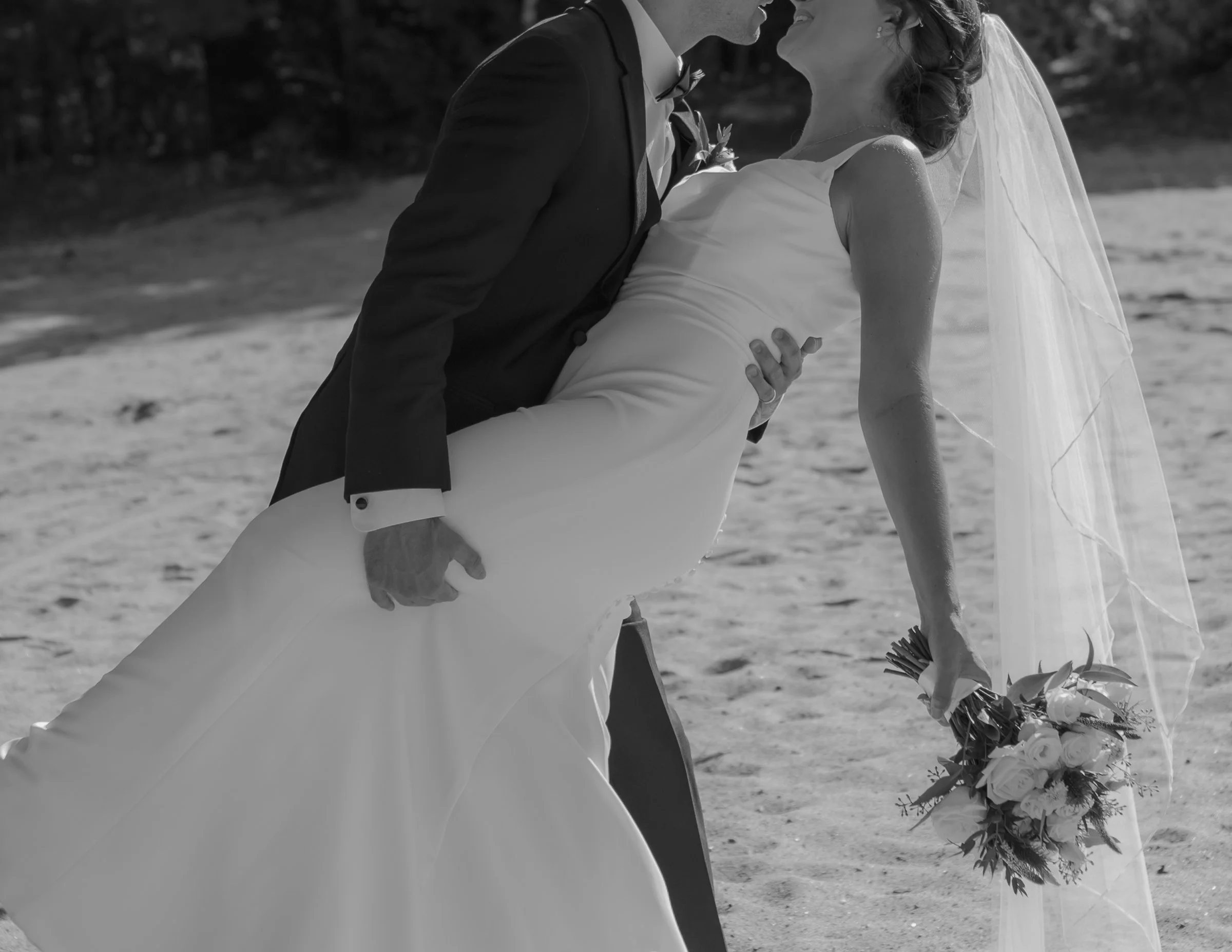 A black and white photo of a bride and groom on a beach. The groom is dipping the bride while she holds a bouquet, and they are about to kiss.