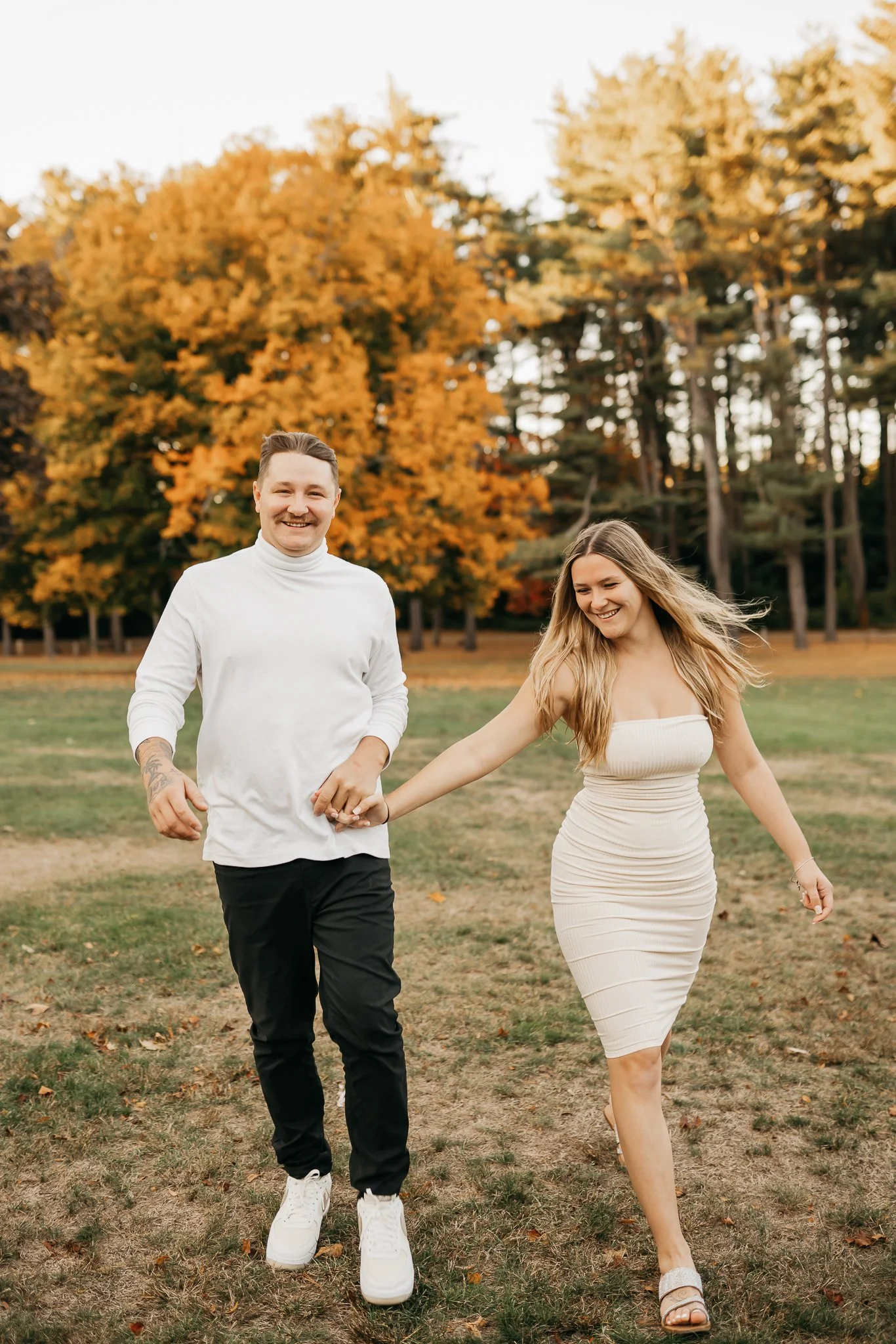 A smiling man and woman holding hands and walking in a park with fall foliage in the background.