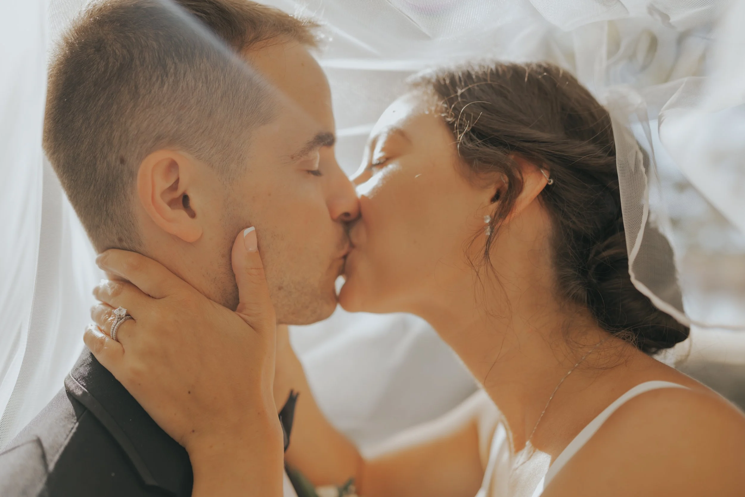A couple sharing an intimate kiss, with the woman holding the man's face and wearing a wedding ring, in a setting with soft natural light.