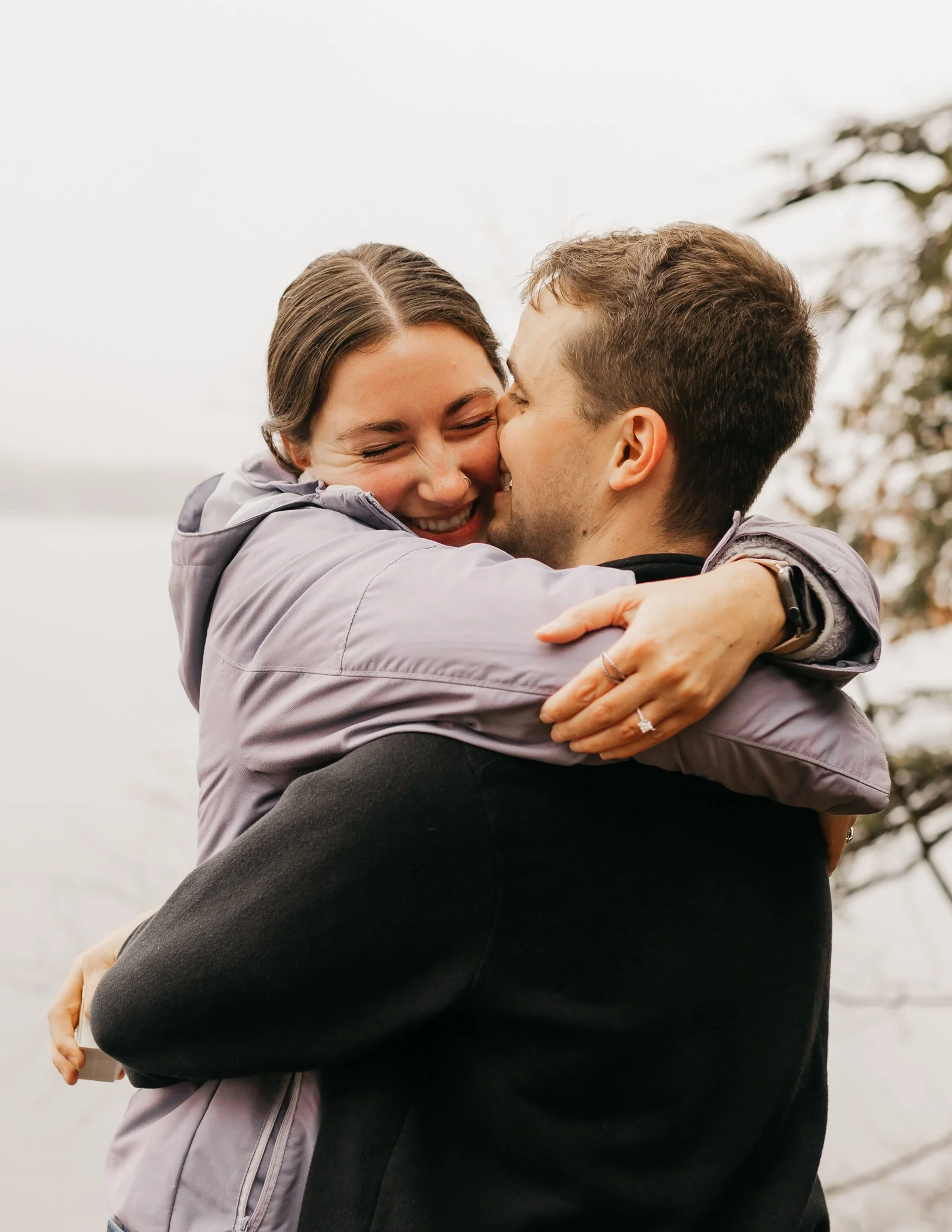 A happy couple hugging outdoors near a body of water, with trees in the background, on a cloudy day.