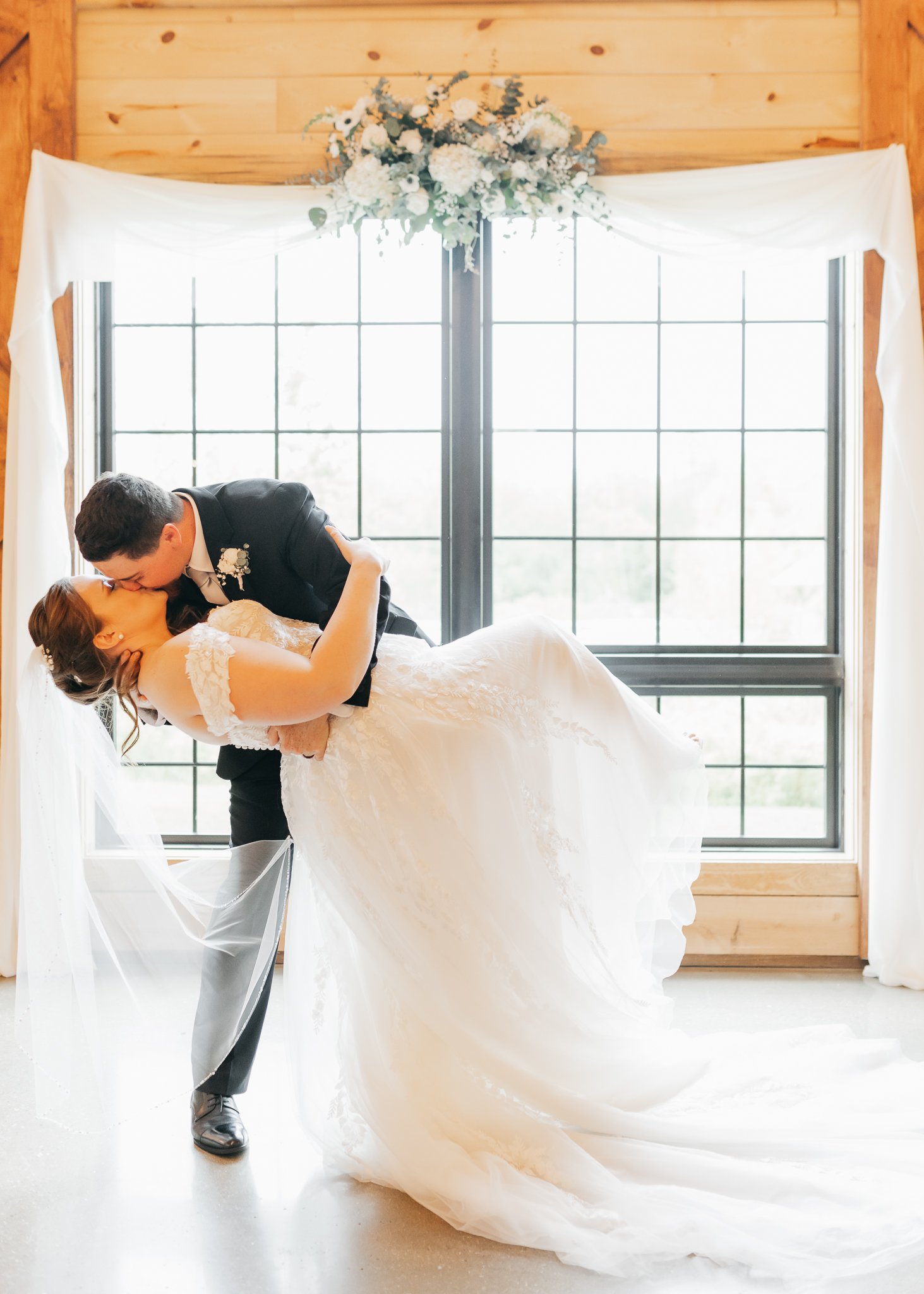 A bride and groom sharing a kiss in front of a large window with white framing, decorated with white flowers and draped white curtains, in a rustic wooden venue.