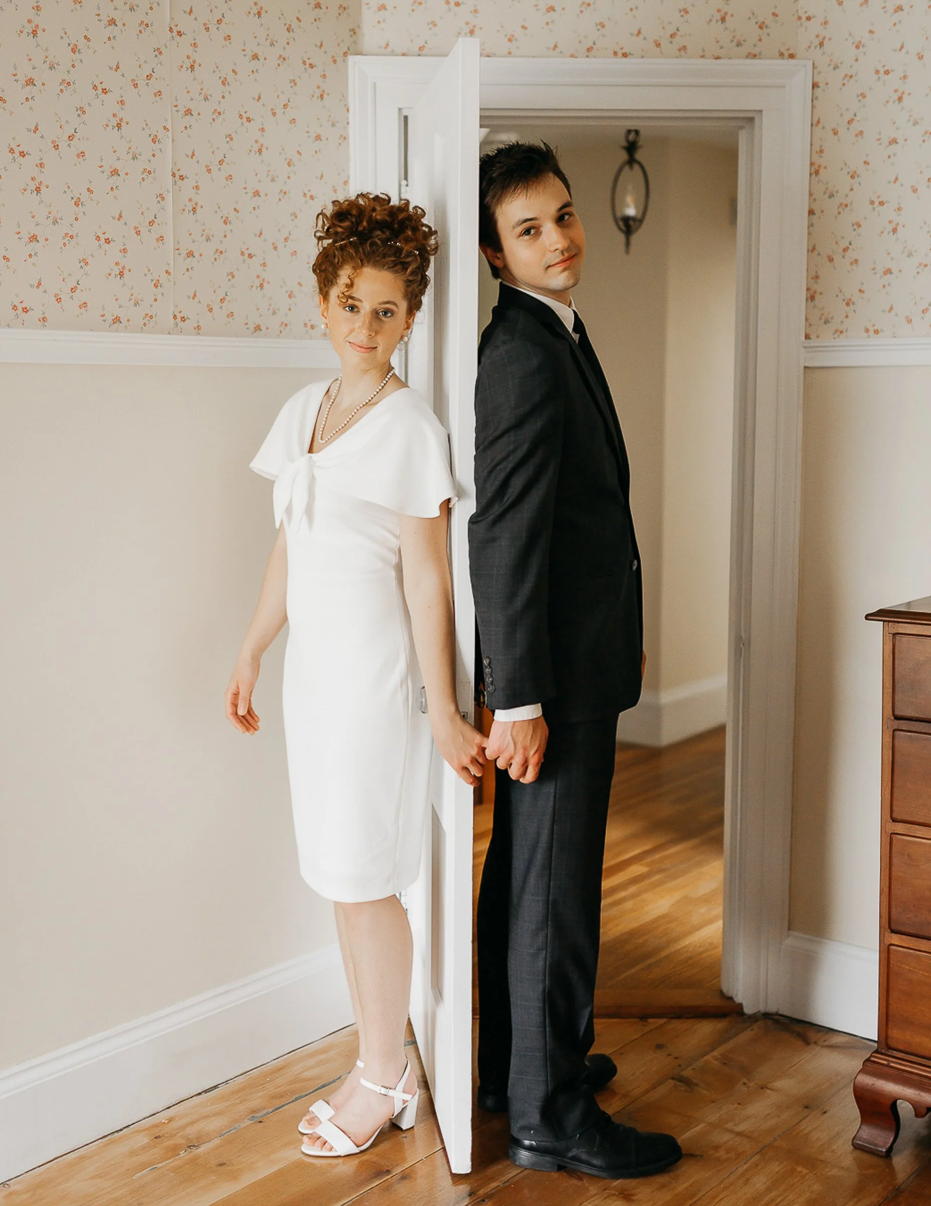 A woman in a white dress and a man in a black suit are standing back to back, holding hands behind a partially open door in a room with floral wallpaper and wooden floor.