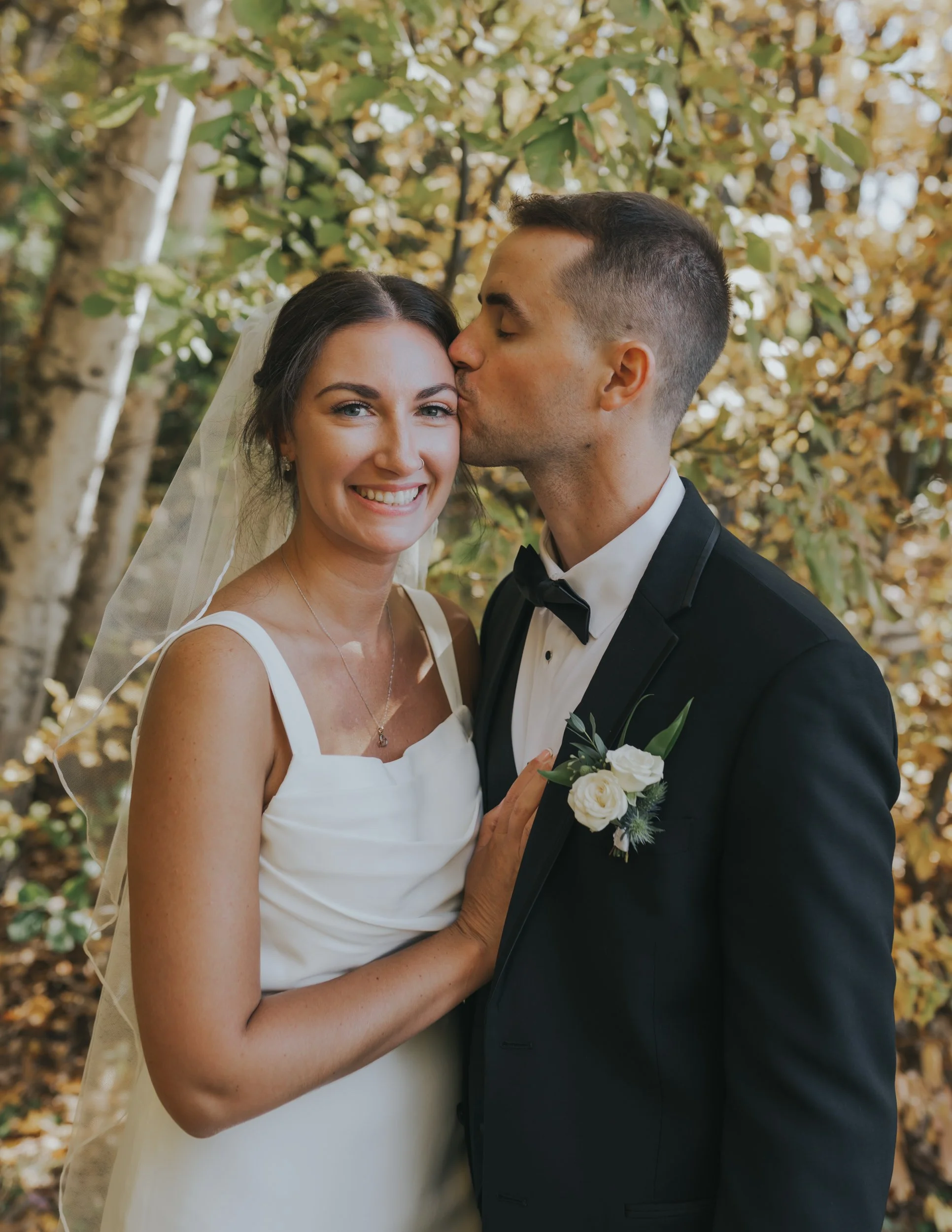 A newlywed couple stands close together outdoors, with the groom kissing the bride on the head. The bride is smiling and wearing a white wedding dress and veil, while the groom is dressed in a black tuxedo with a bow tie and white boutonniere. The ba