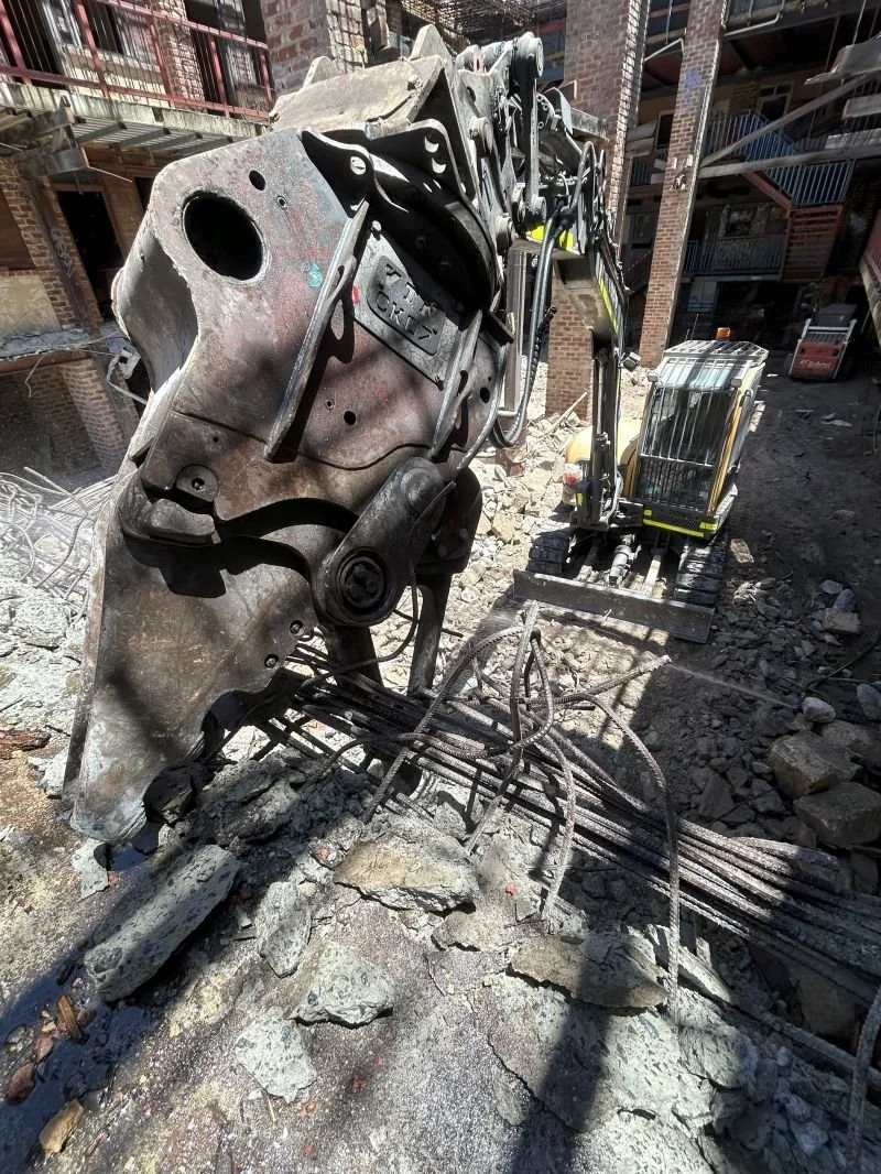 Close-up of a construction excavator on a building site with broken concrete and rebar. The excavator arm is visible, and the background shows brick building and scaffolding.