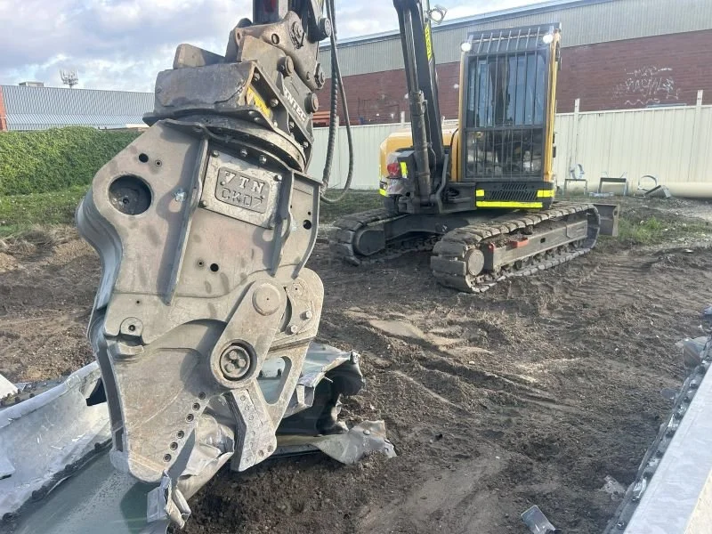 Construction site with a small excavator and a mechanical arm or attachment for an excavator, dirt ground, and a building in the background.