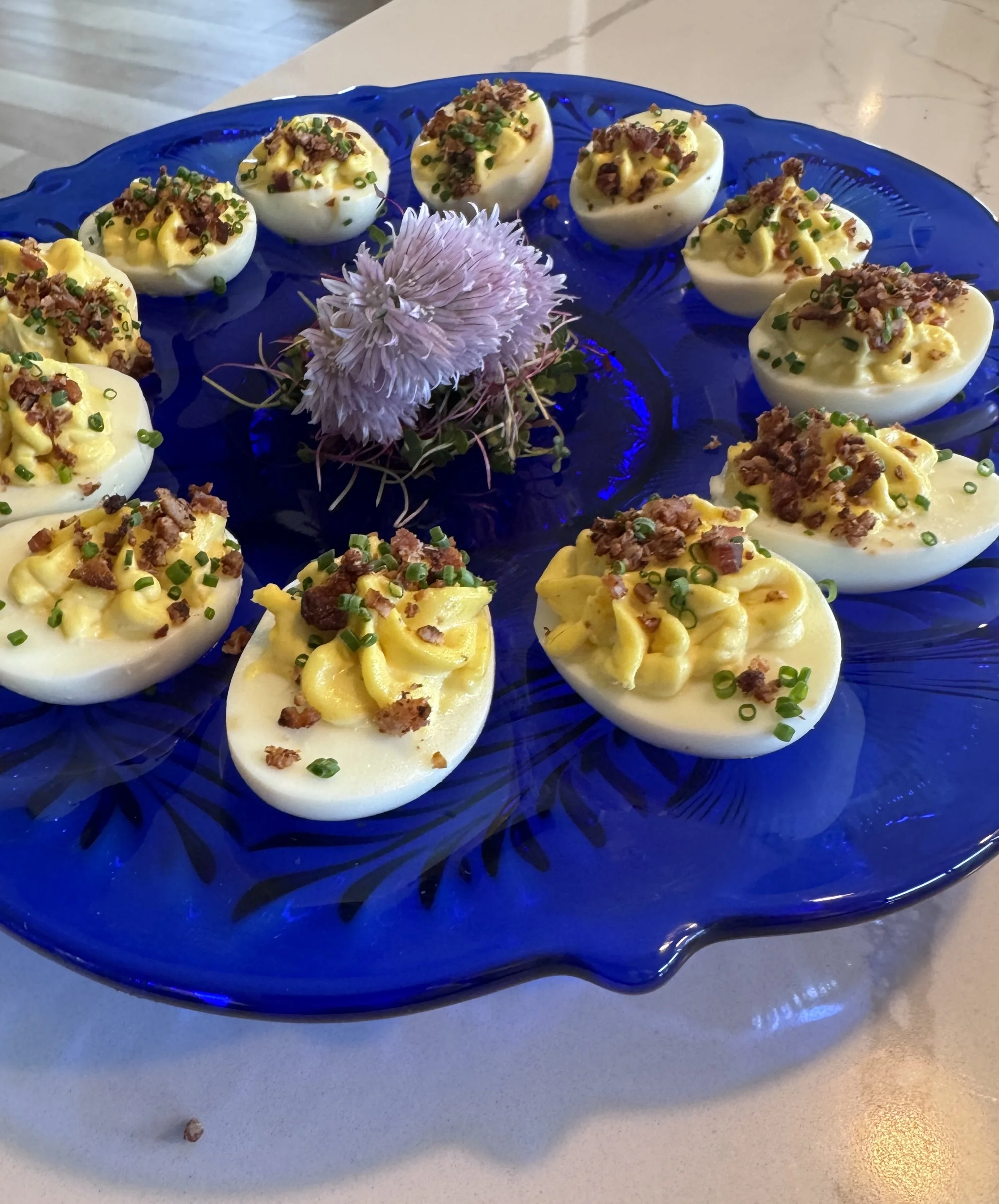 Halved hard-boiled eggs topped with chopped bacon, chives, and mustard, arranged in a circle on a blue glass serving platter with a garnish of a fresh chive flower from the garden.