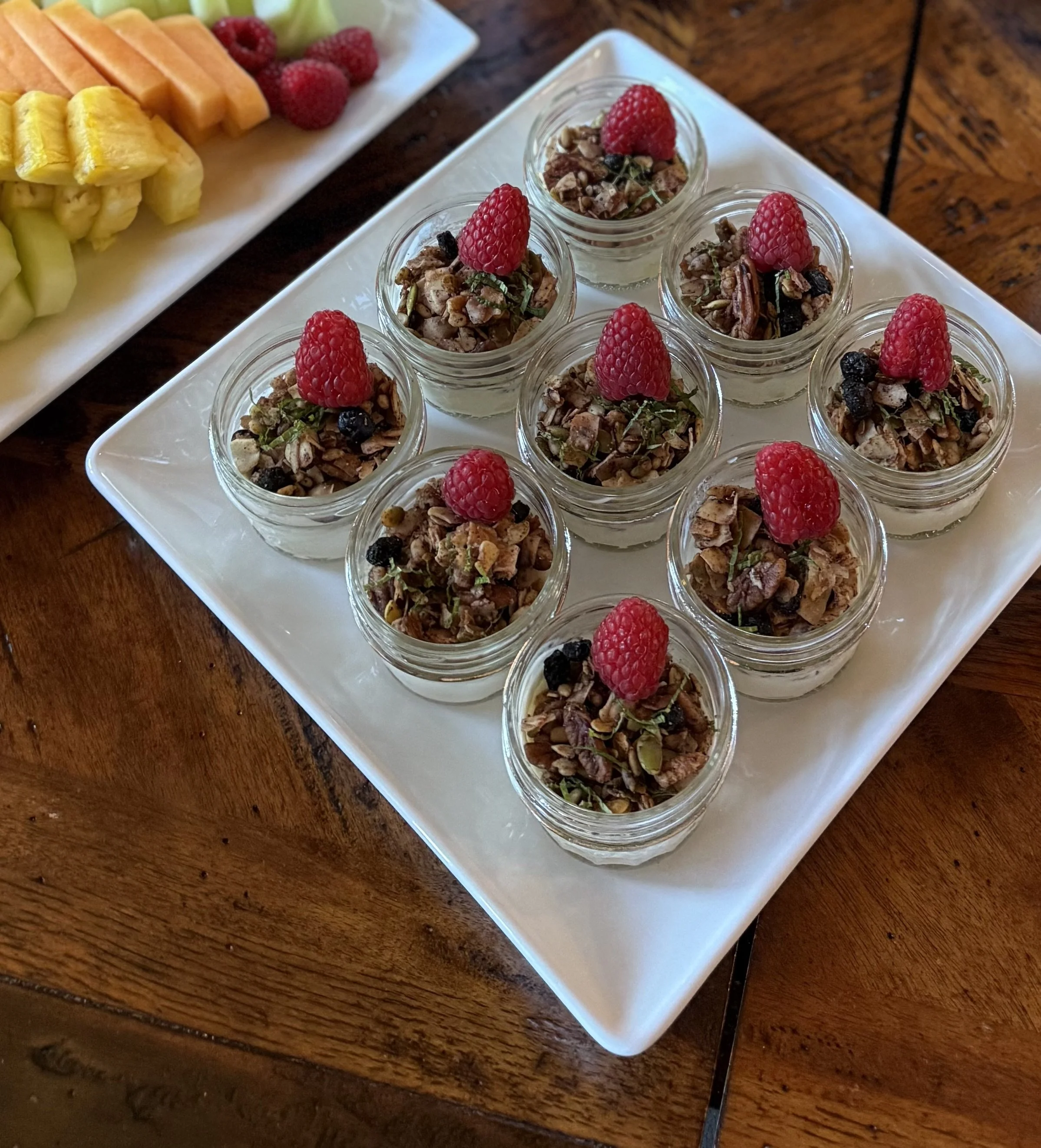 Mini mason jar yogurt parfait filled with layered granola, topped with a raspberry, on a white rectangular platter.
