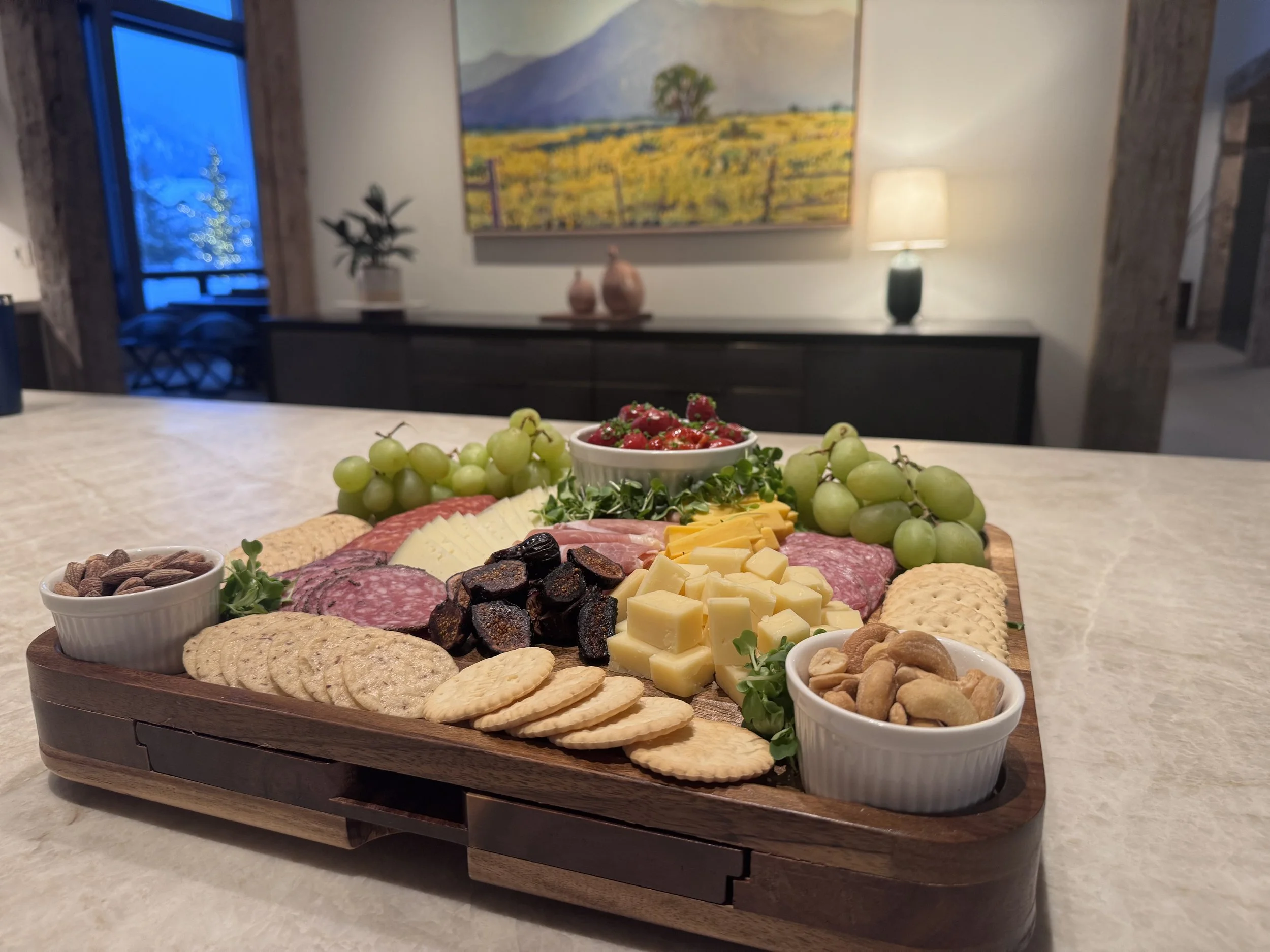 A wooden cheese and charcuterie board featuring grapes, crackers, cheese cubes, sliced meats, dried figs, nuts, and a bowl of mixed nuts, set on a kitchen counter with a modern interior in the background.