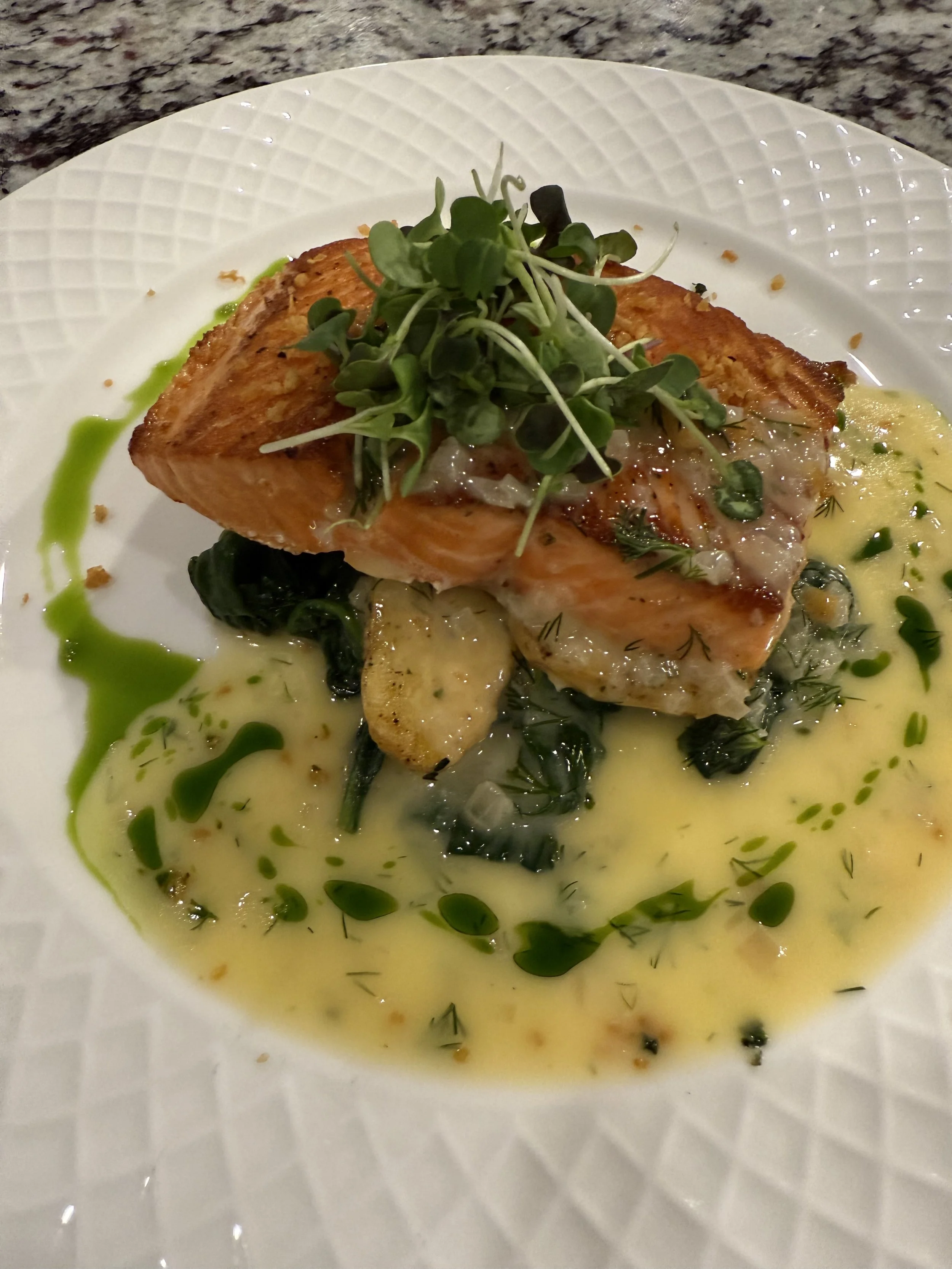 Pan-seared trout with beurre blanc, basil oil, micro greens, and roasted potatoes on a white textured plate.