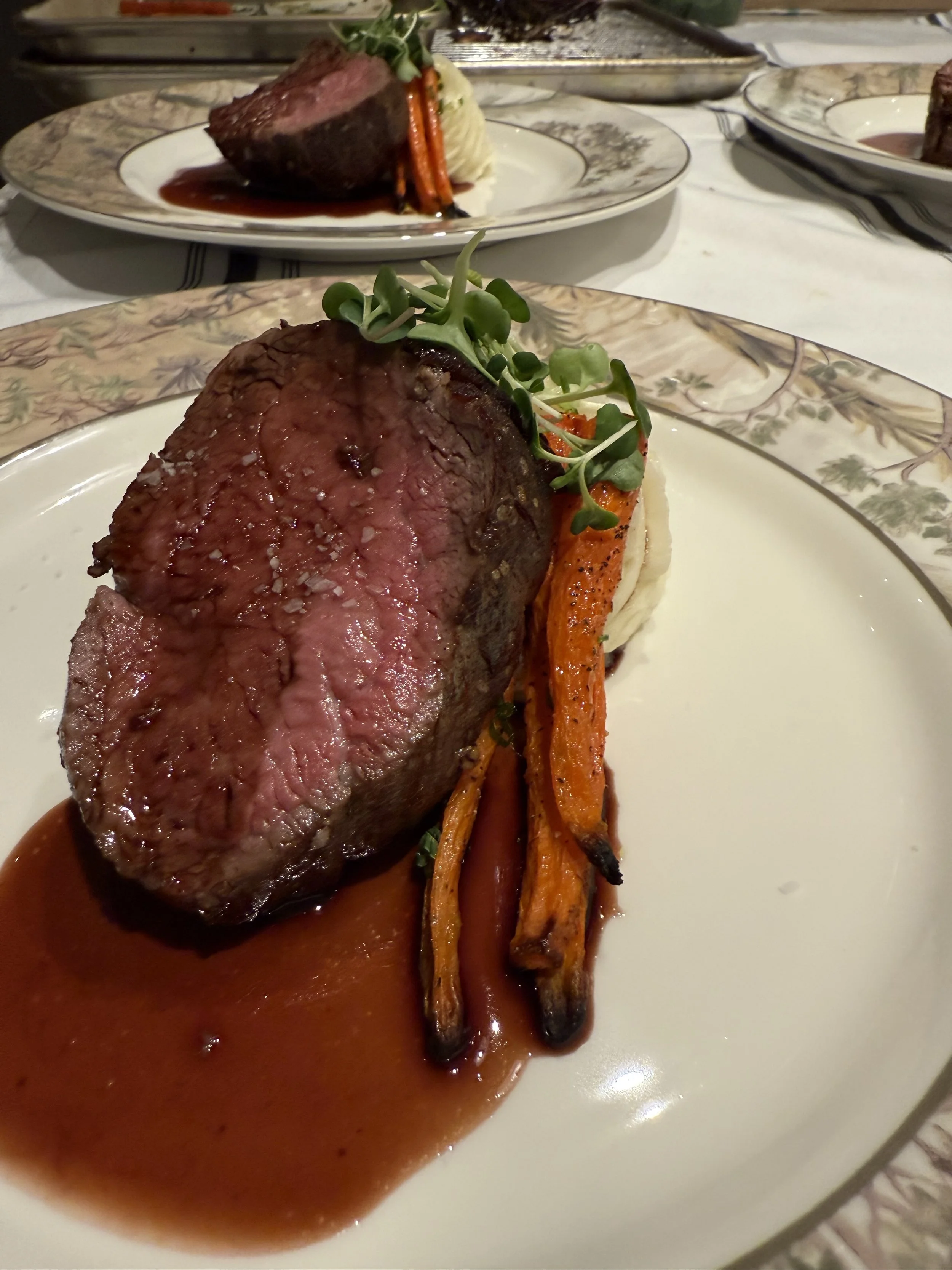 A plate with a medium-rare beef steak, roasted carrots, mashed potatoes, and microgreens, served with a brown sauce.