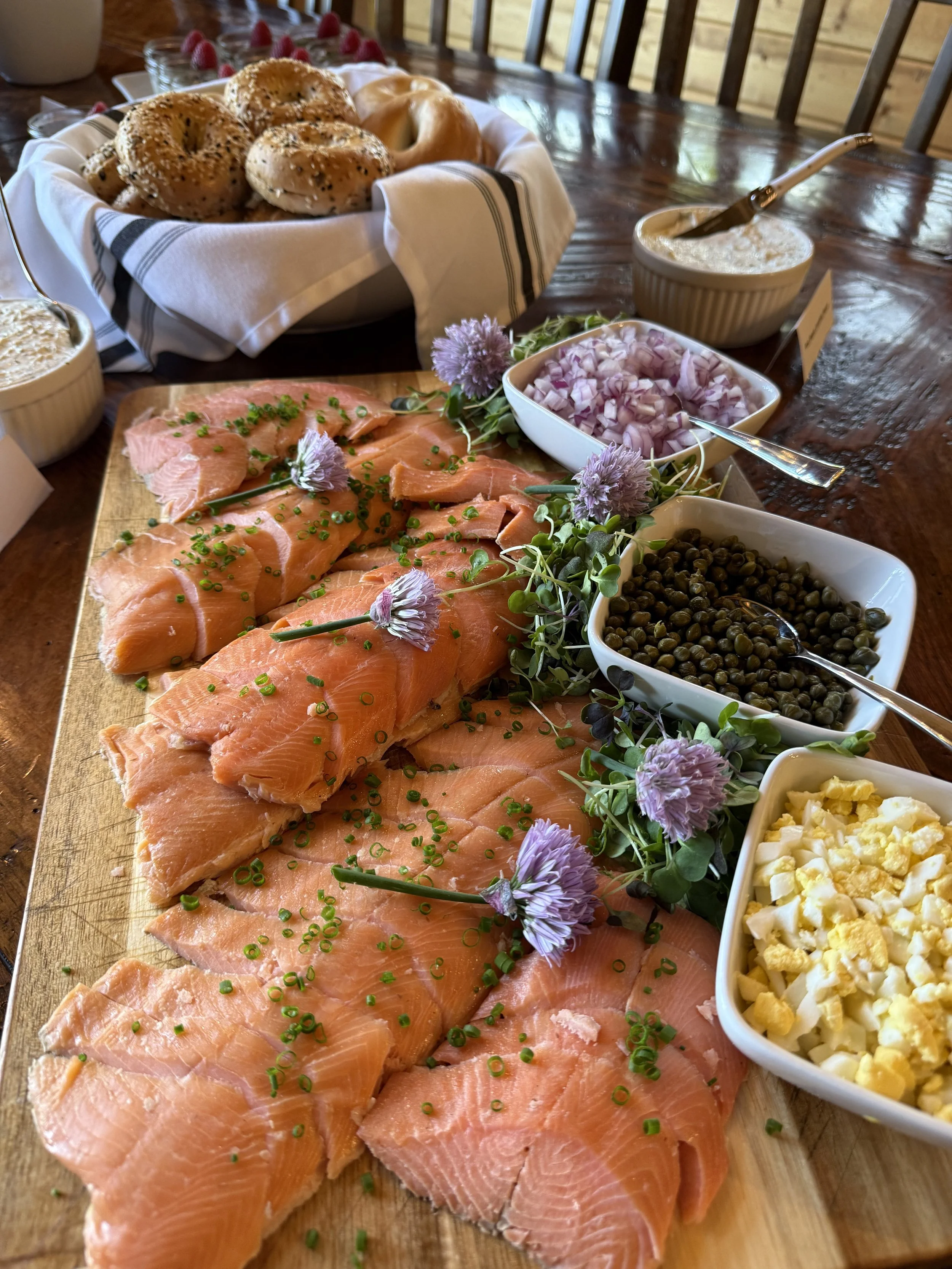 Smoked trout slices garnished with chopped chives on a wooden platter accompanied by bowls of chopped onions, capers, chopped hard-boiled eggs, cream cheese, with a basket of assorted bagels in the background.
