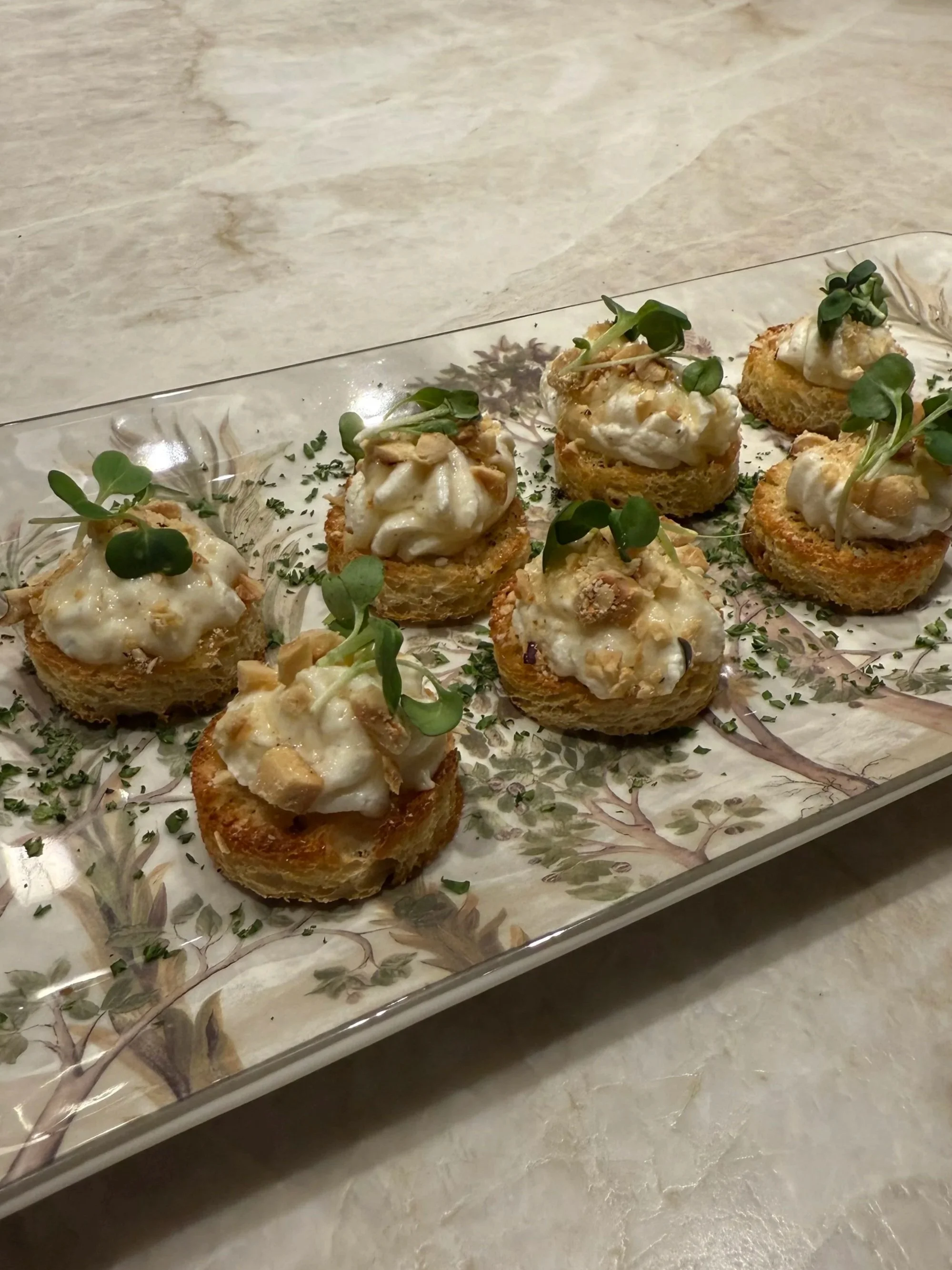 Eight toasted bread rounds topped with whipped ricotta cheese, chopped nuts, and garnished with micro greens on a decorative rectangular glass tray.