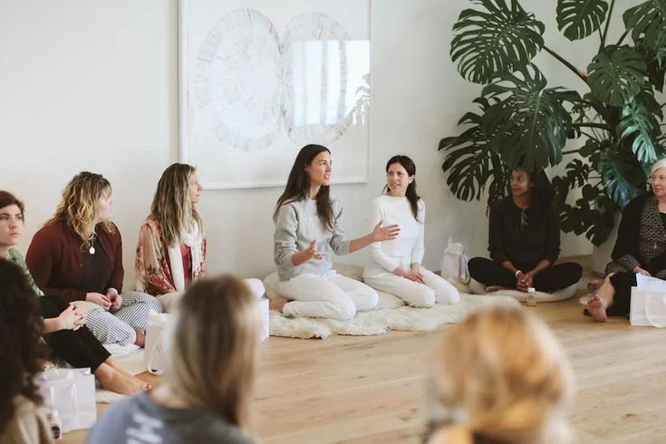 Group of women sitting on the floor in a circle during a discussion or workshop in a bright room with large plants and artwork on the wall.