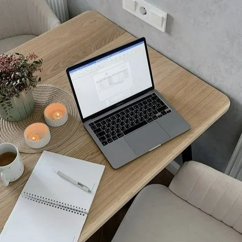 Wooden desk with an open laptop displaying a spreadsheet, a notebook with a pen, a white mug, two lit candles in white holders, and a vase with flowers.