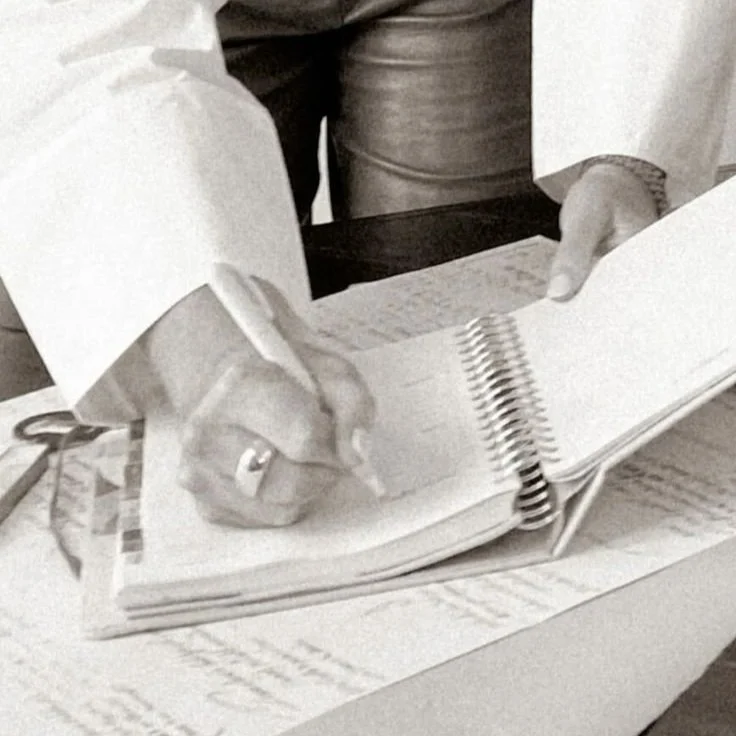 Person wearing a long-sleeve shirt writing in a spiral notebook with a pen on a table.