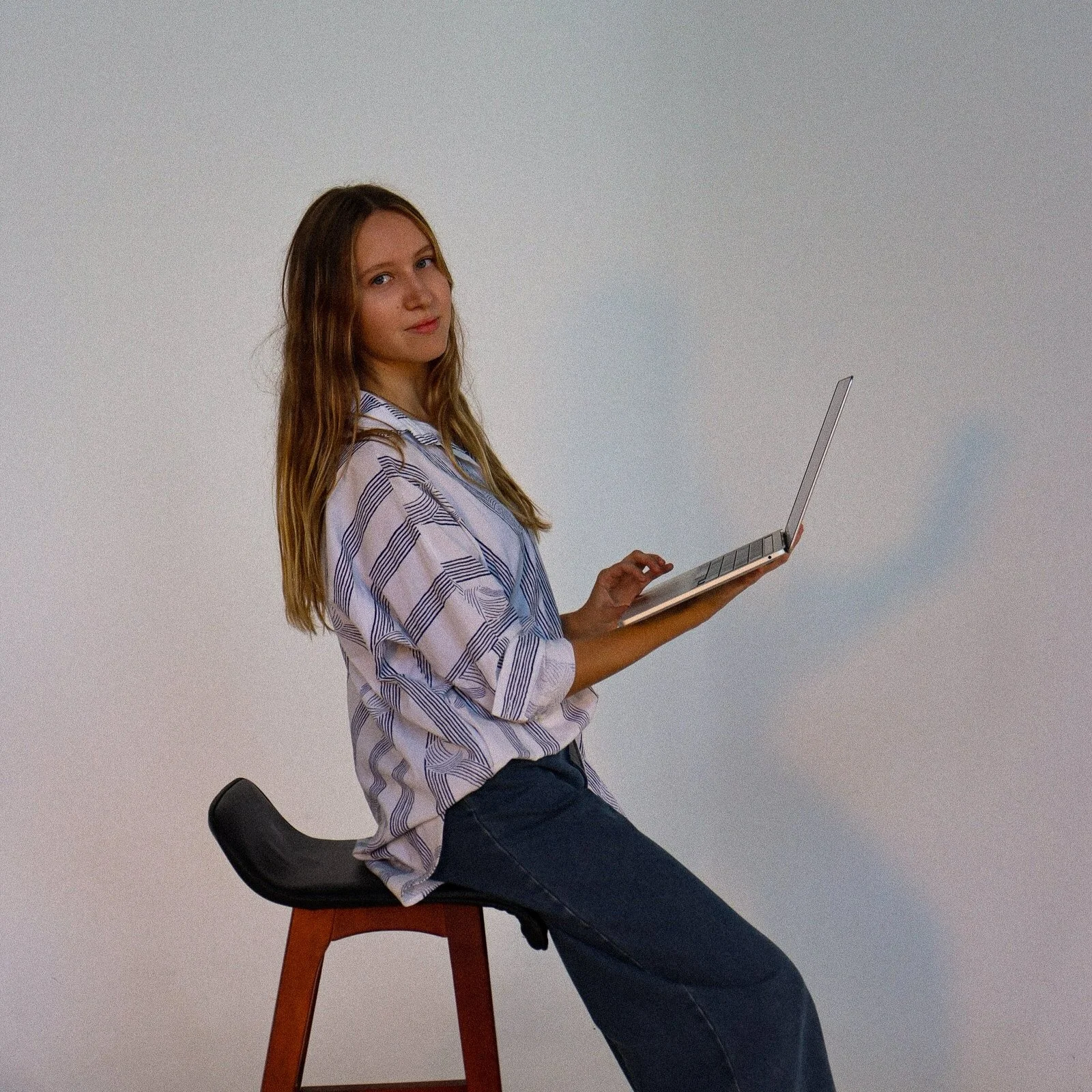 A young woman with long wavy brown hair sitting on a black and brown wooden stool, wearing a loose striped button-up shirt and dark jeans, holding an open laptop and looking at the camera.