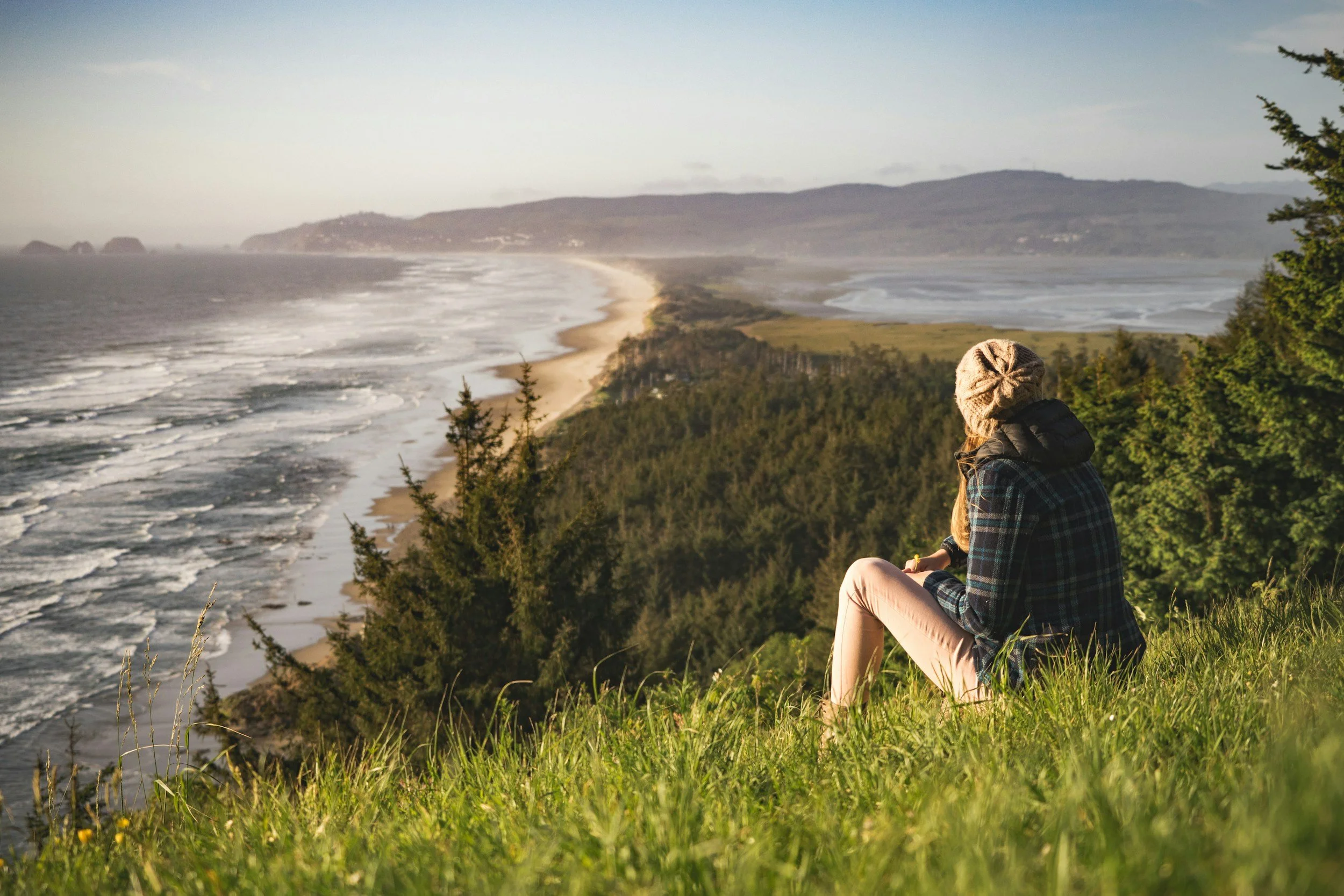 A woman sitting on a grassy hill overlooking the ocean and a sandy beach at sunset, surrounded by trees.