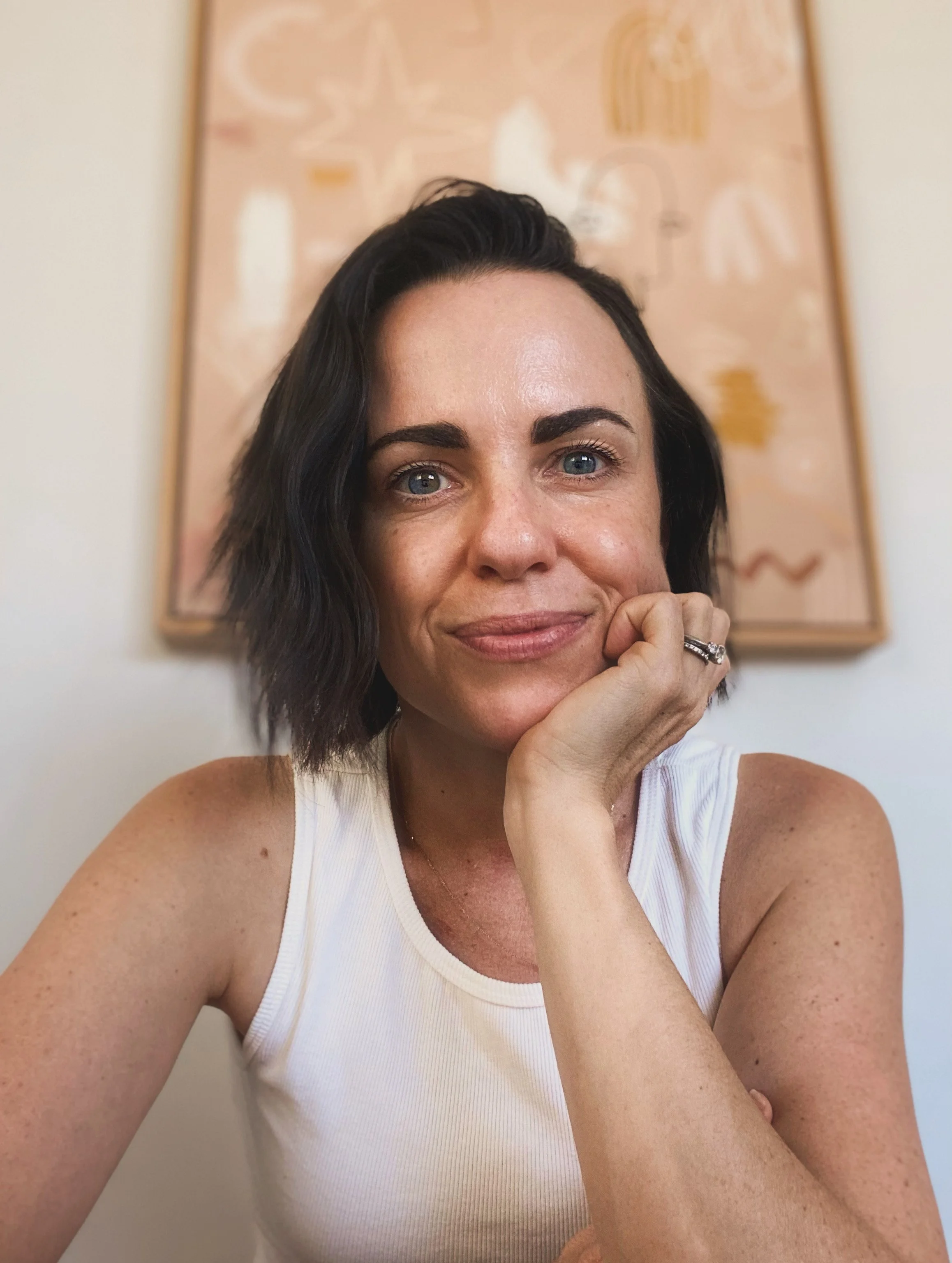 A woman with short dark hair and blue eyes, wearing a white sleeveless top, resting her chin on her hand, smiling softly, with a pink and beige abstract art background.