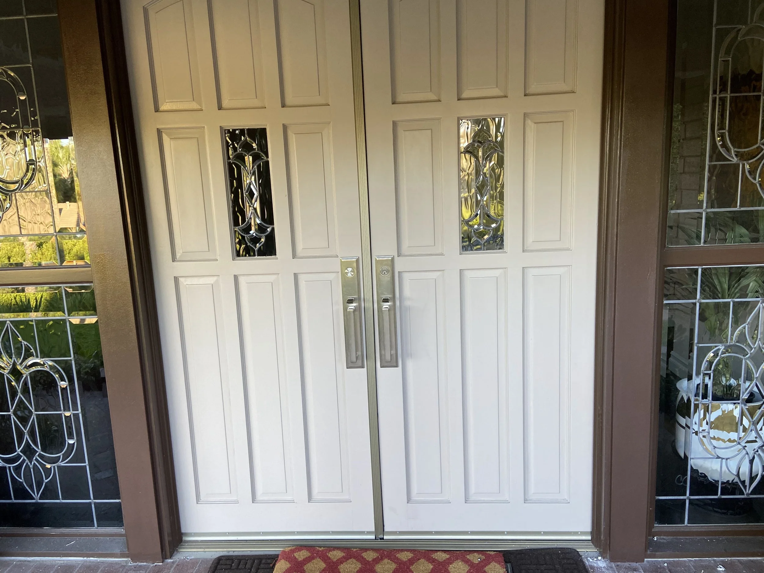Closed white front door with decorative glass panels and brown trim, flanked by glass sidelight panels with ornamental metal designs, and a red patterned doormat in front.