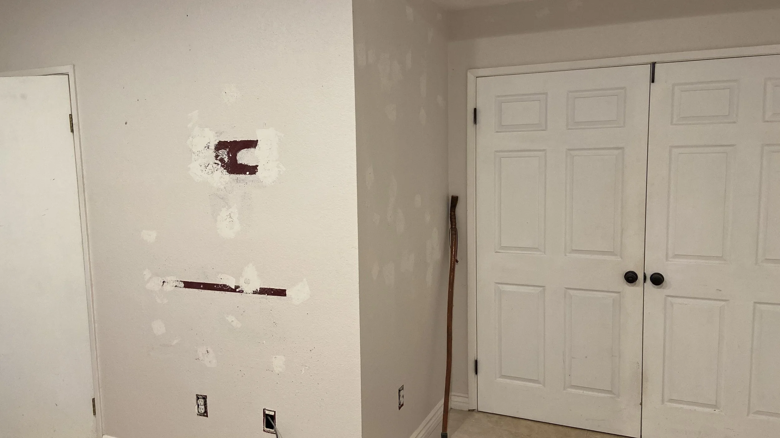 Interior wall under renovation with patches of drywall mud, a wooden walking stick, and a set of white double closet doors.