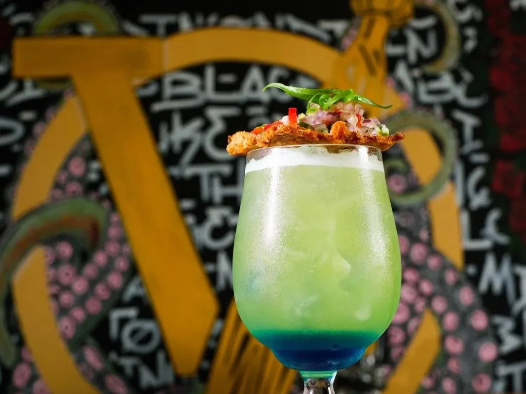 Colorful cocktail in a tall glass with layered colors of blue, green, and yellow, garnished with a lime wedge, a crispy chip, and herbs, against a vibrant black background with yellow and pink patterns.