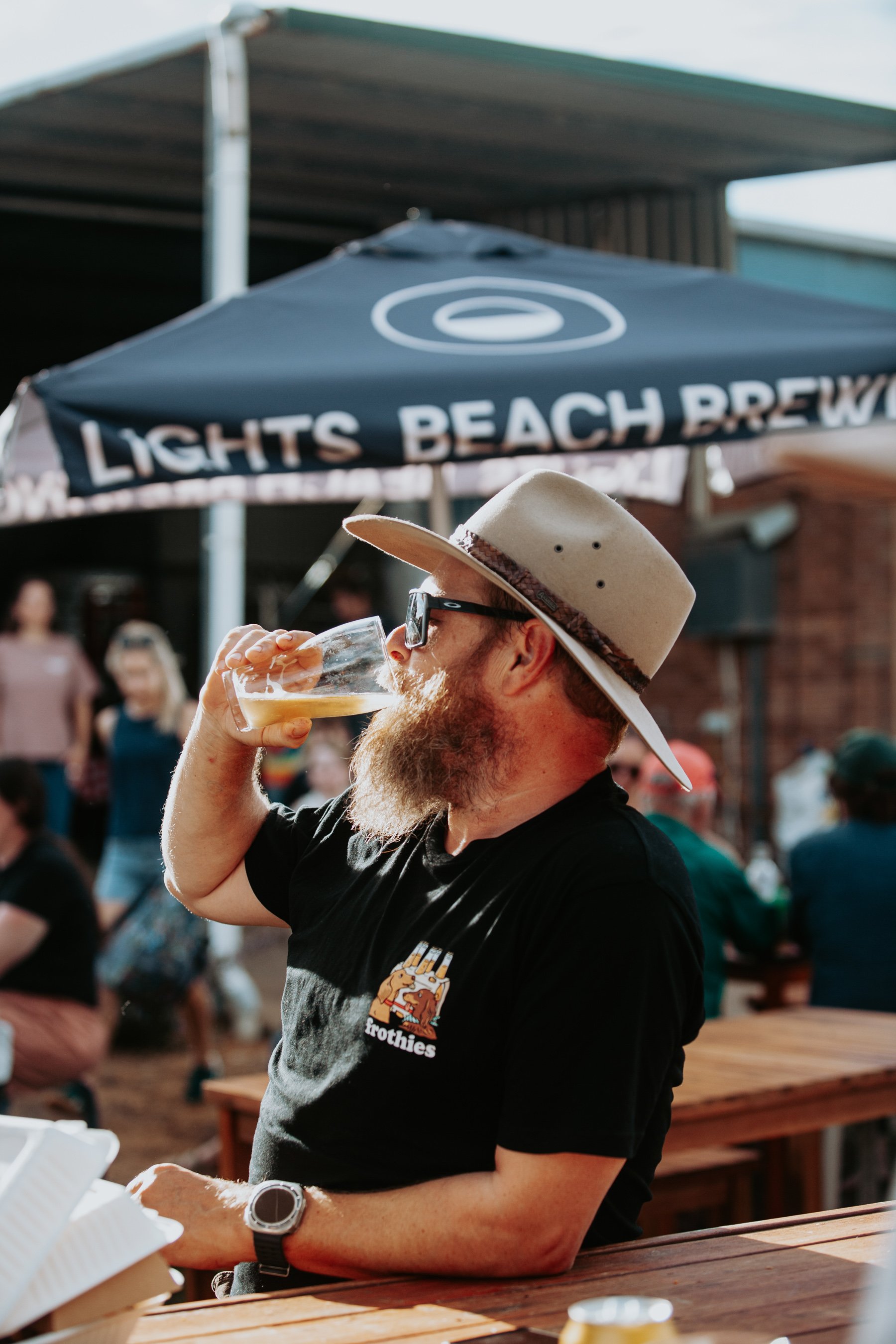 A man in an Akubra hat drinking a beer out of a glass in a sunny beergarden with people in the background