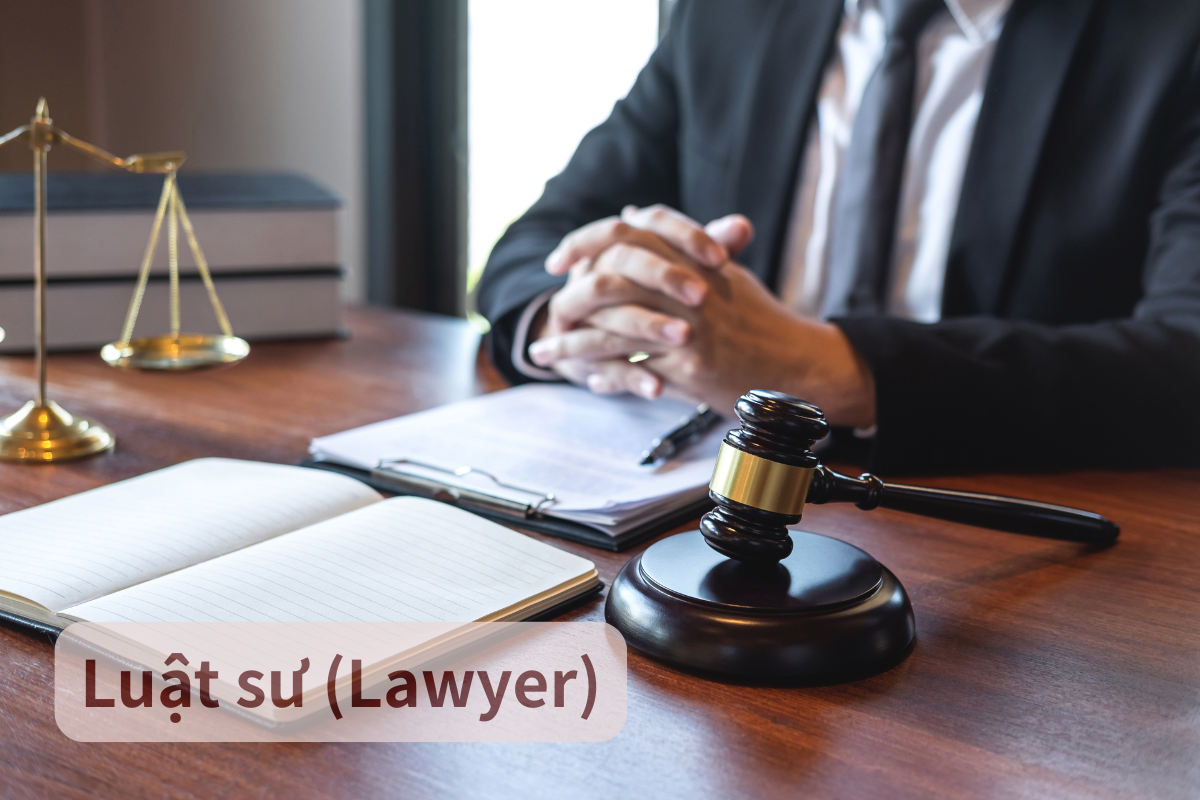 A lawyer sitting at a wooden desk, with a gavel, legal books, open notebook, and pen on the desk, dressed in a dark suit with hands clasped. The image indicates a legal or courtroom setting.