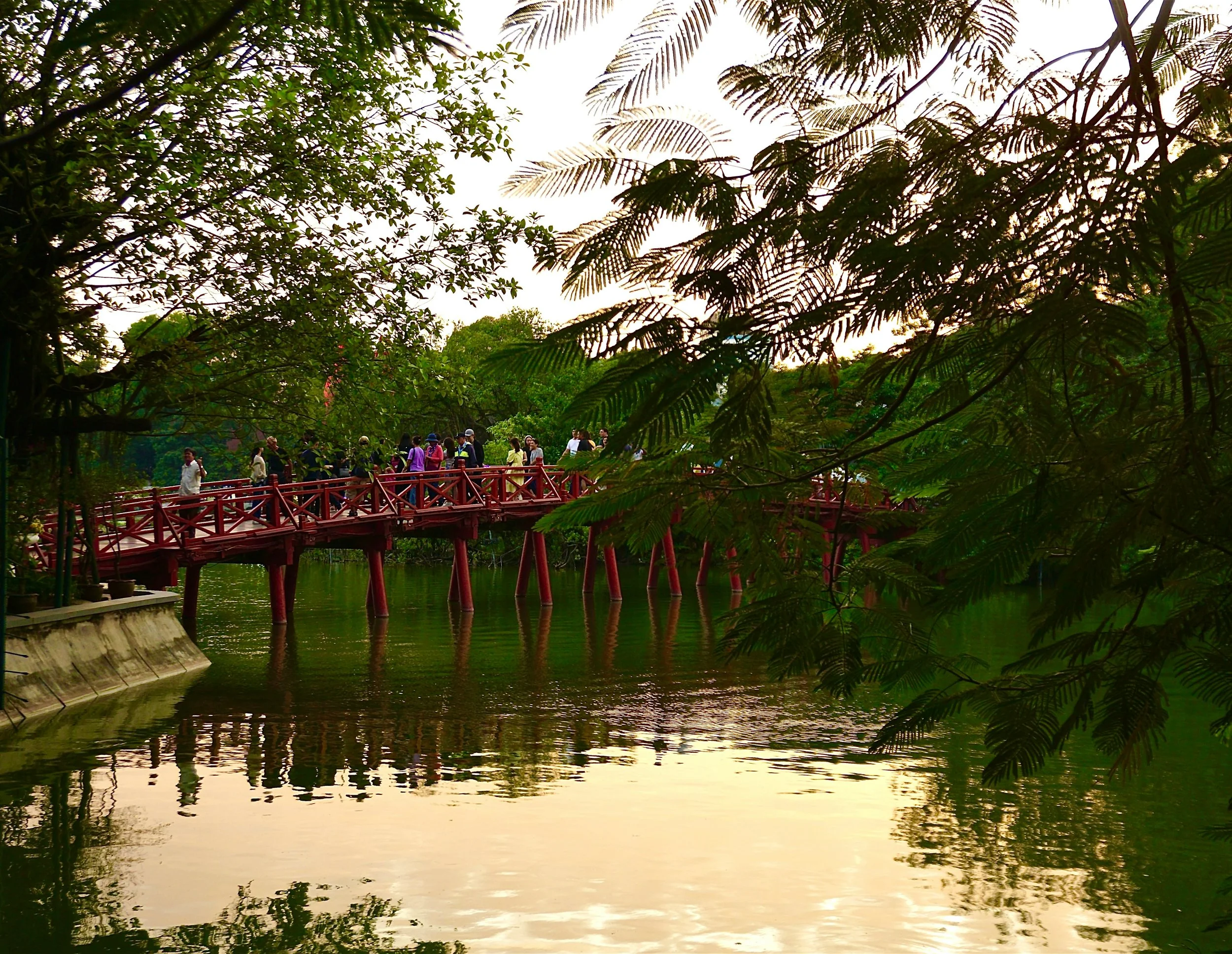 Red bridge over a calm lake with people walking, surrounded by lush green trees and reflected in the water.