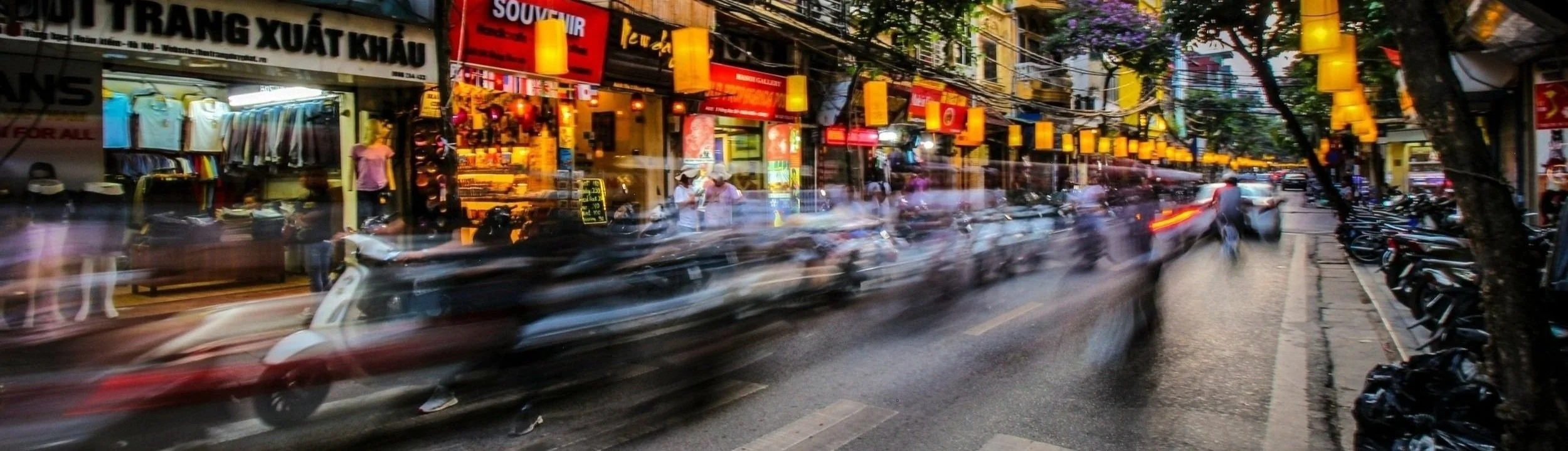 Busy street scene in Vietnam with shops, blurred motion of scooters, and lit lanterns.