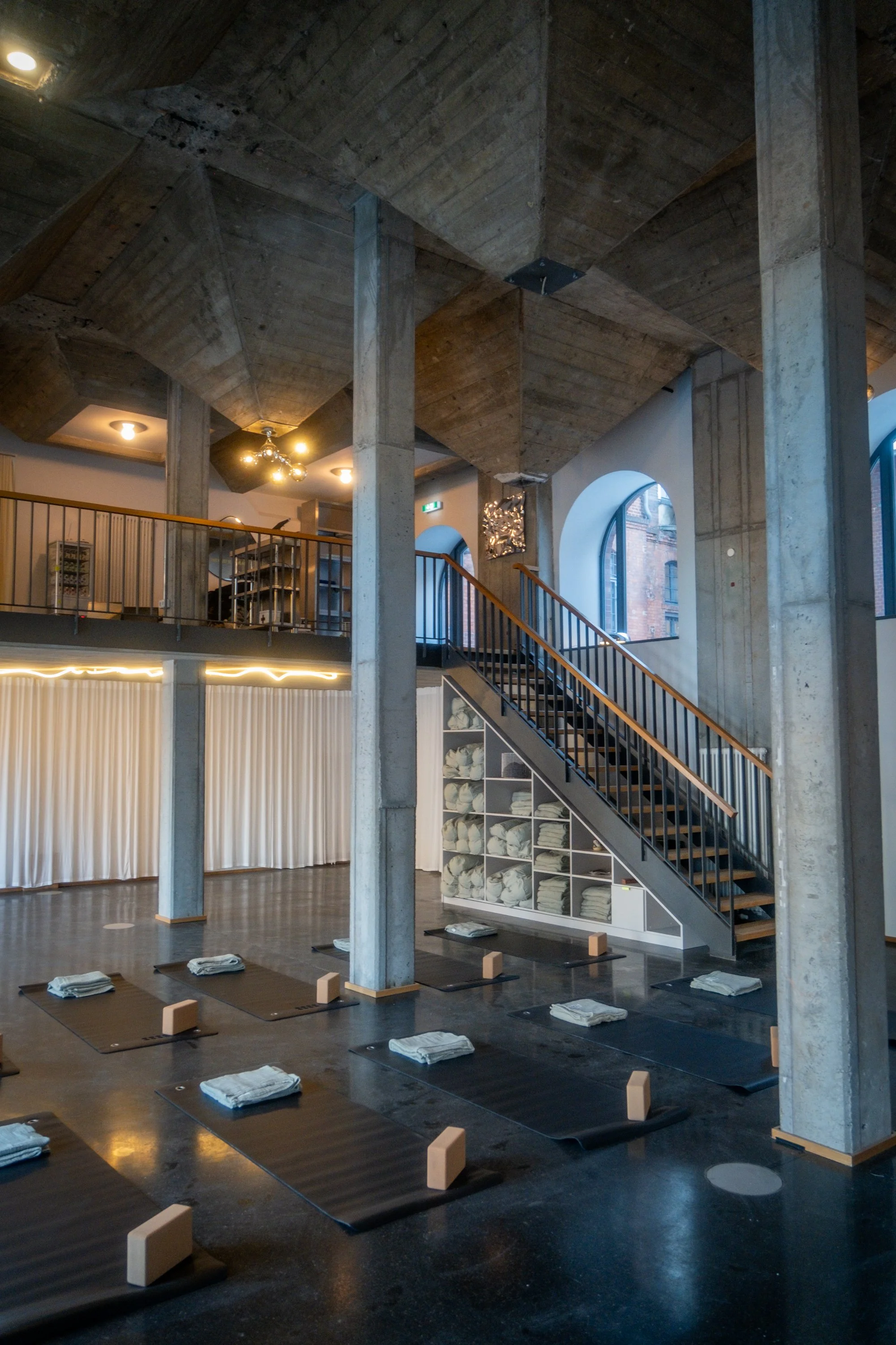 Yoga class studio with mats, towels, and blocks arranged on the floor, upper and lower levels, exposed concrete columns and ceiling, wooden staircase, large arched windows, and soft lighting.