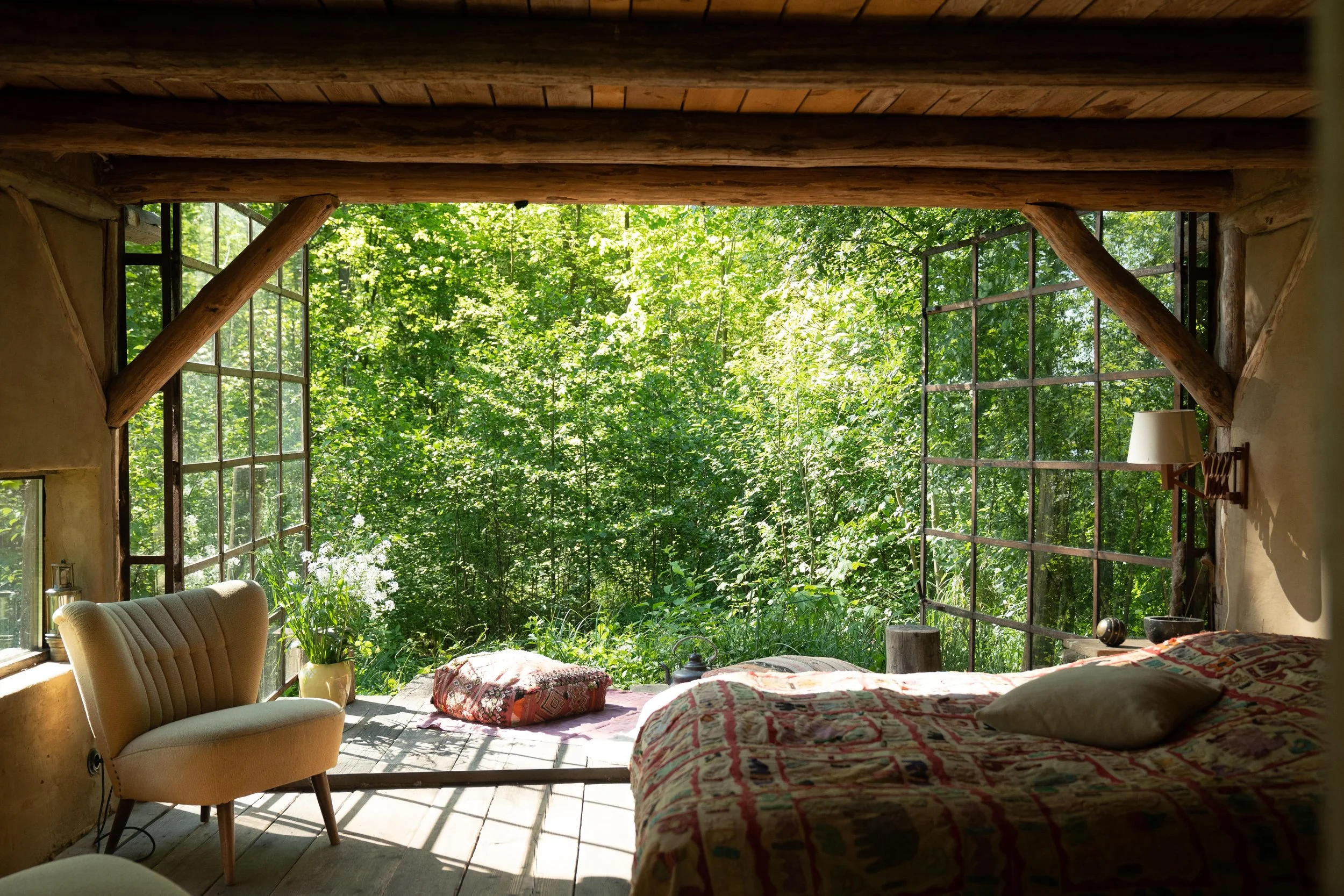 A cozy bedroom with a large open window overlooking a lush green forest, natural wooden beams, a bed with patterned bedding, a beige armchair, a potted plant, and soft sunlight illuminating the space.