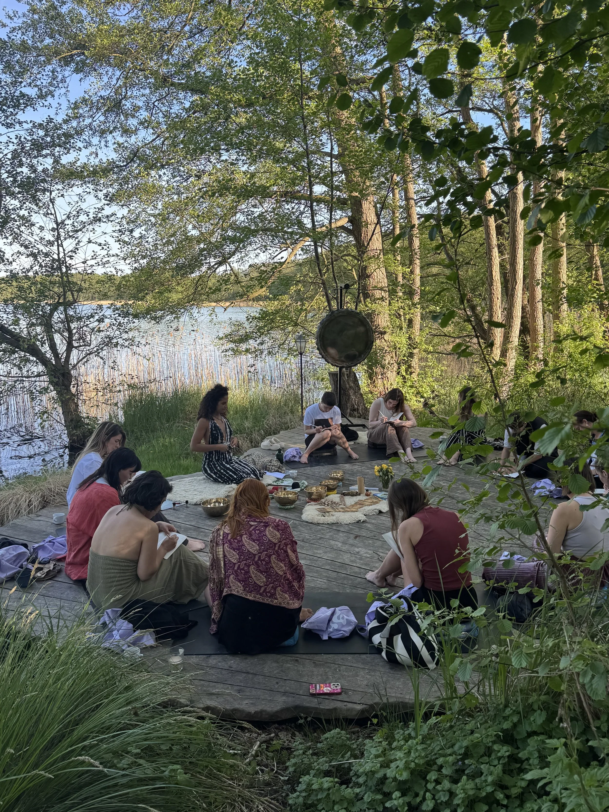 A group of people practicing meditation outdoors on a wooden platform near a lake surrounded by trees, with their eyes closed or looking down, sitting cross-legged or on their knees, with some items placed in the center such as bowls and flowers.