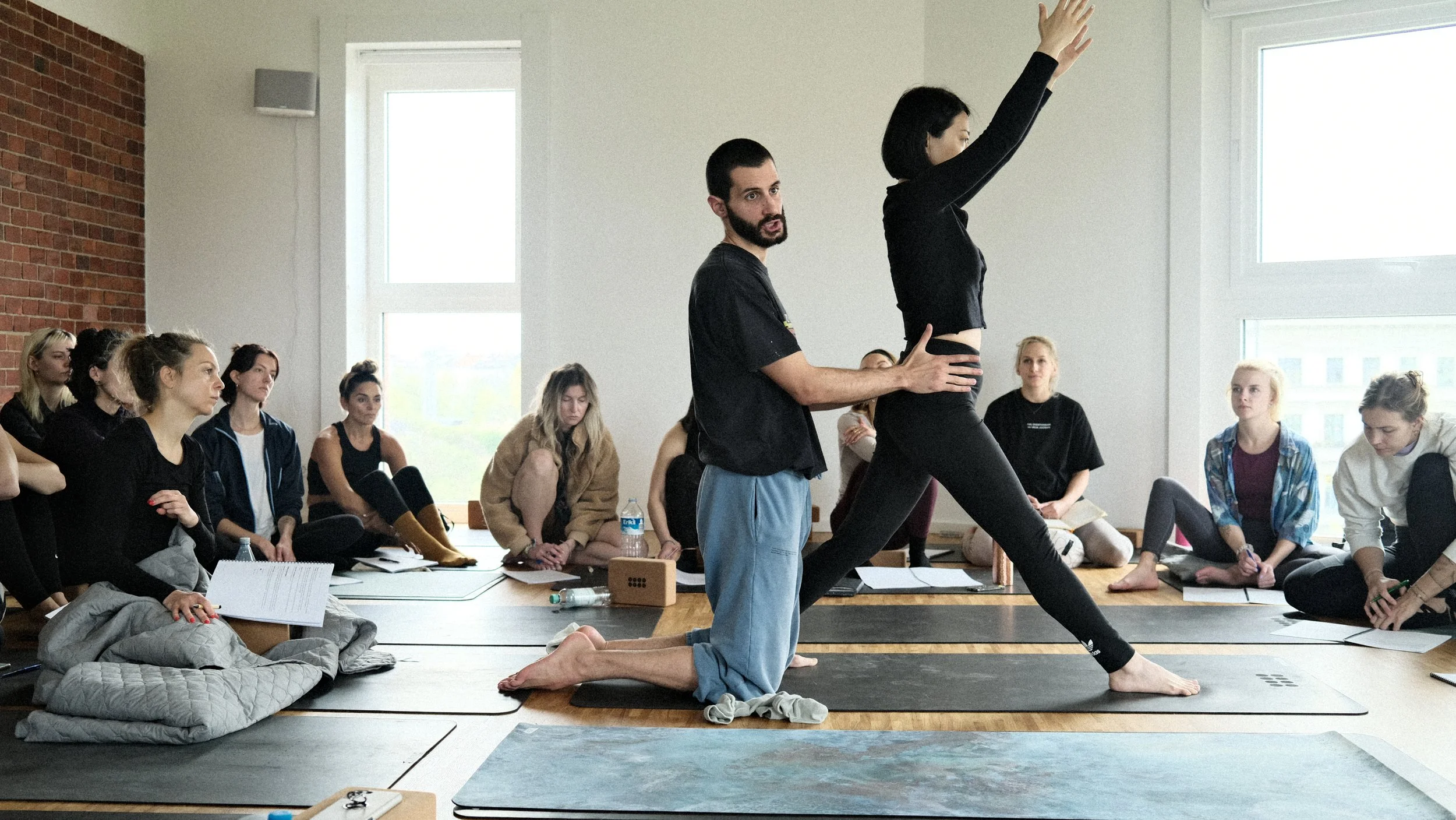 A group of women participating in a yoga or fitness class, sitting on mats in a spacious room with large windows. A man is guiding a woman in a pose where she is kneeling and stretching her arms upwards, while the others watch or prepare for their turns.
