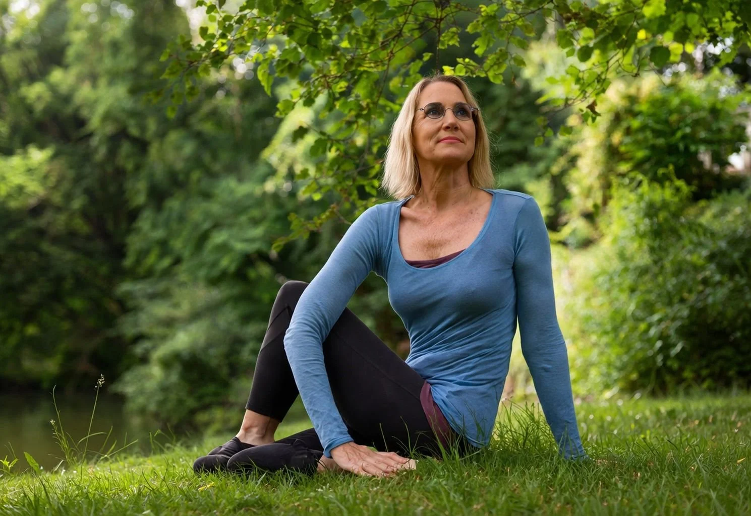 A woman doing yoga outdoors on a grassy area surrounded by trees.
