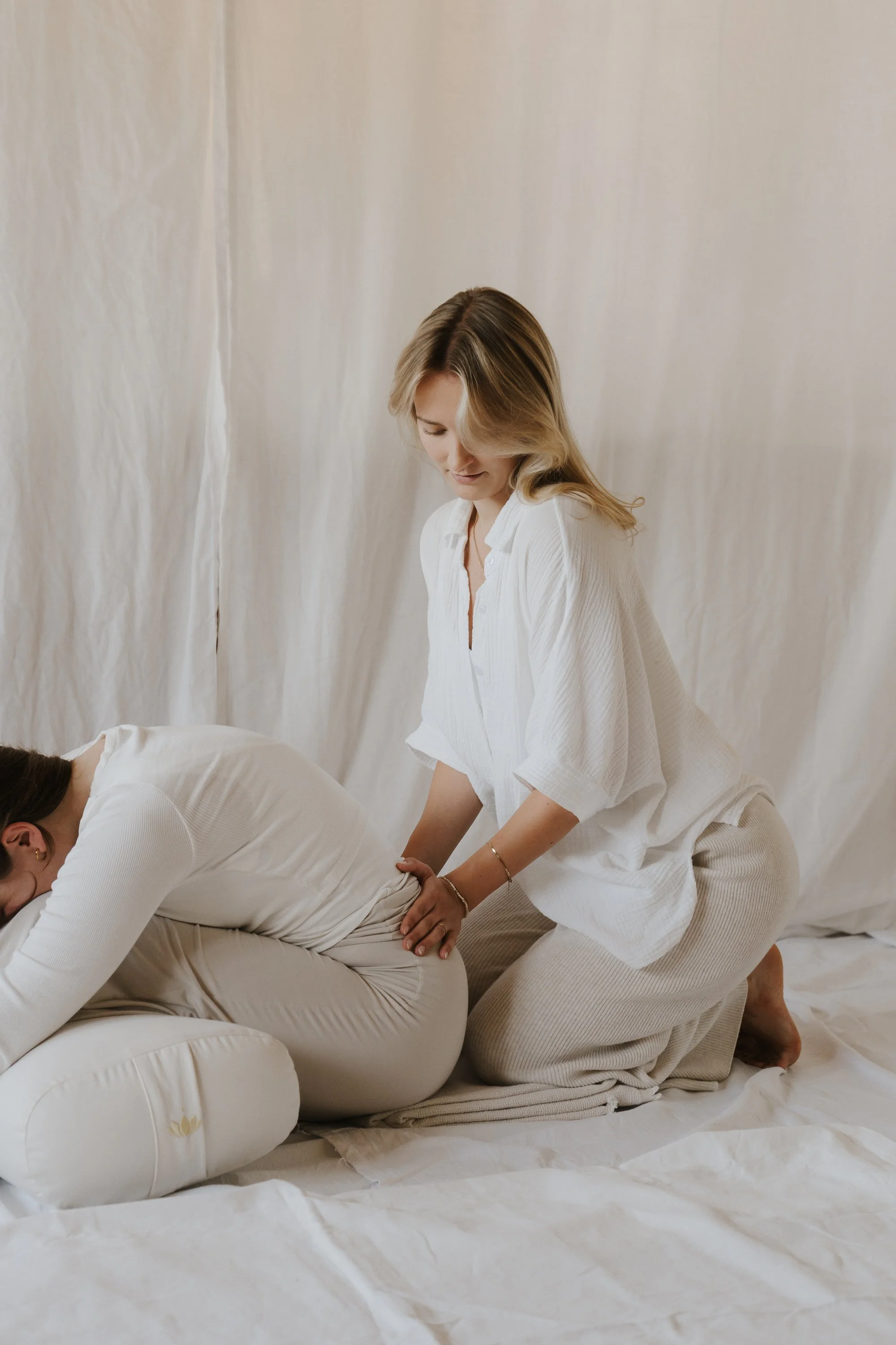 A woman is giving a massage to a person lying on the floor, both dressed in white from the waist up, in a room with white curtains or a backdrop.