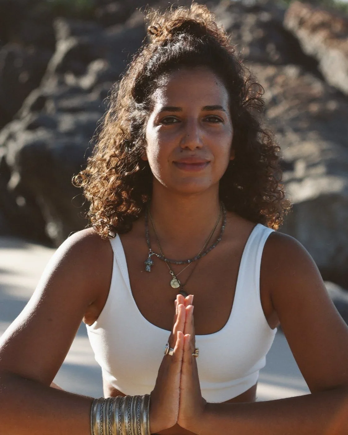A woman with curly hair practicing yoga outdoors, standing with palms pressed together in prayer pose, wearing a white tank top and layered necklaces, with rocks and sunlight in the background.