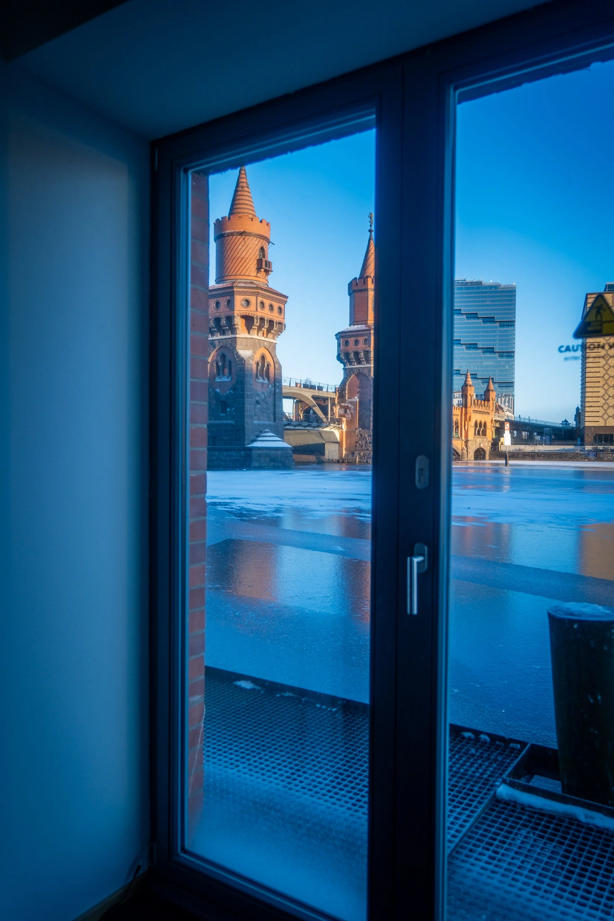 View of the Oberbaum Bridge in Berlin, Germany, seen through a glass door, with frozen water and modern buildings in the background.