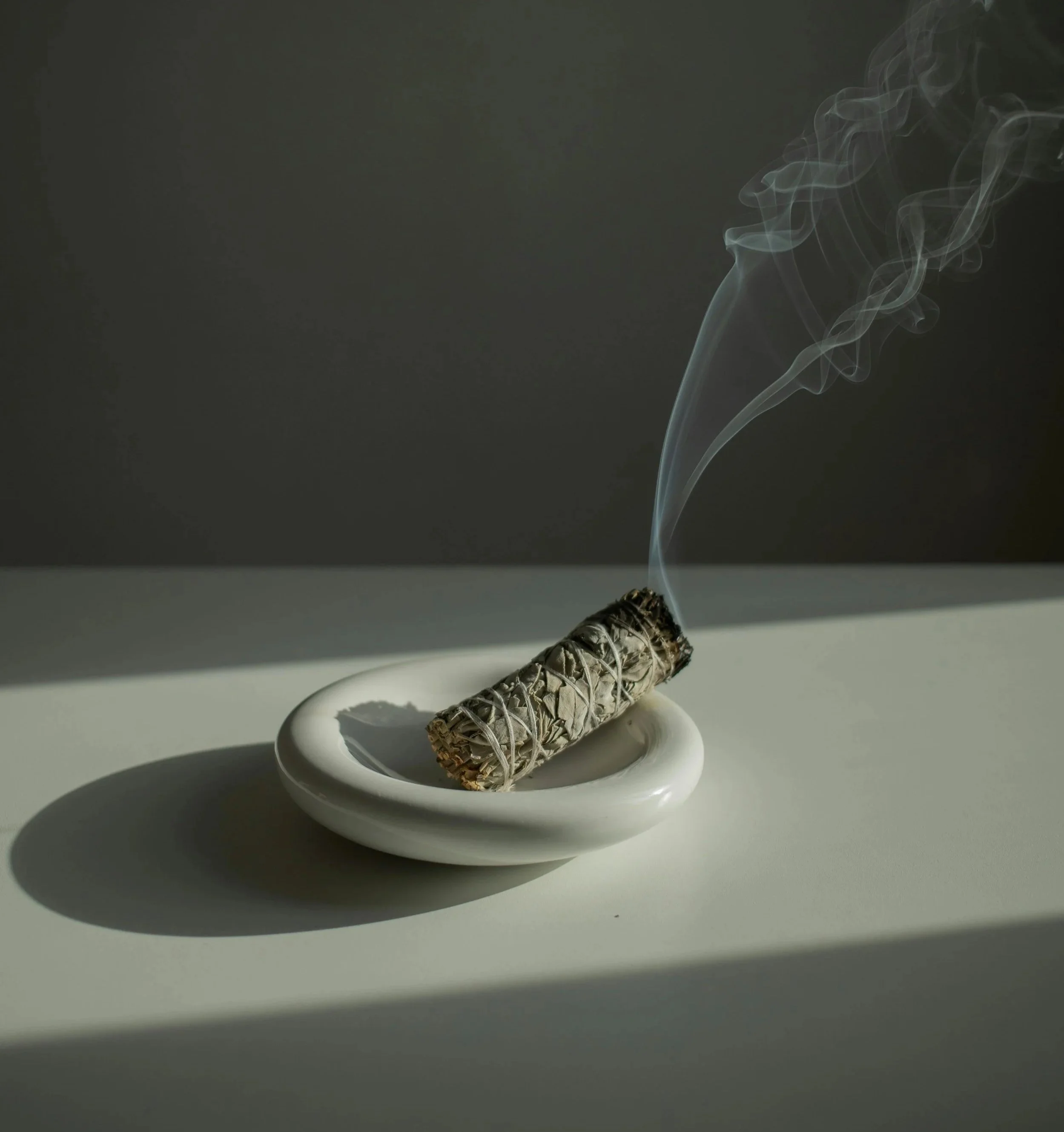 Burned sage bundle resting on a white ashtray with smoke curling upward, casting shadows on a gray surface and background.