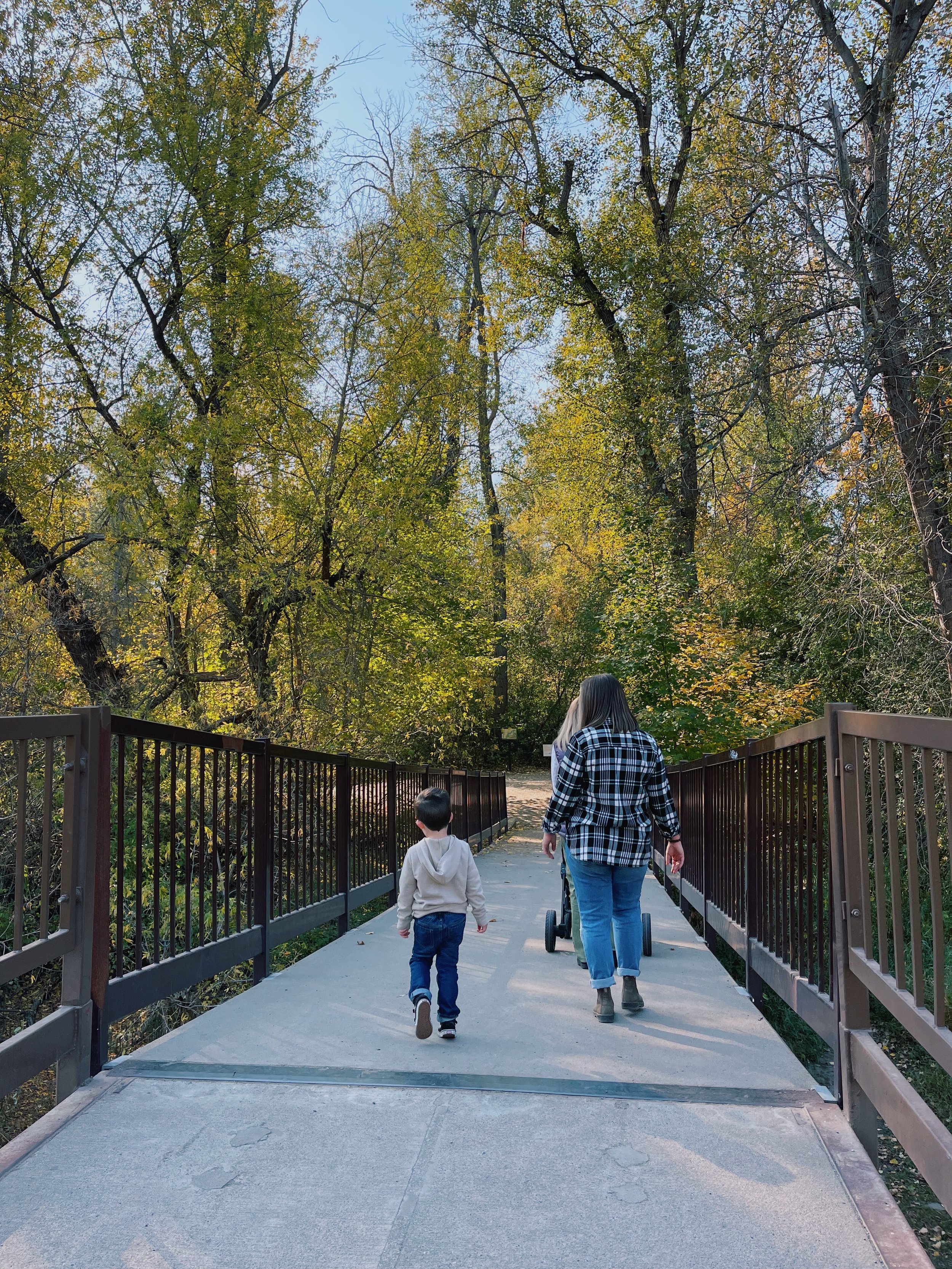 A woman and a young boy walk along a paved trail in a wooded park. The woman is pushing a stroller and wearing a black and white checkered shirt with blue jeans. The boy, dressed in a light gray hoodie and blue jeans, walks ahead. Tall trees with green and yellow leaves line the trail, indicating autumn.