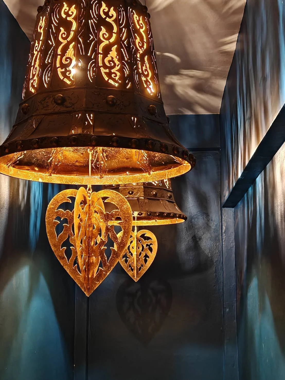 An ornate hanging lantern with carved metal design, emitting a warm yellow glow, and decorated with three heart-shaped cutouts with bat designs inside.