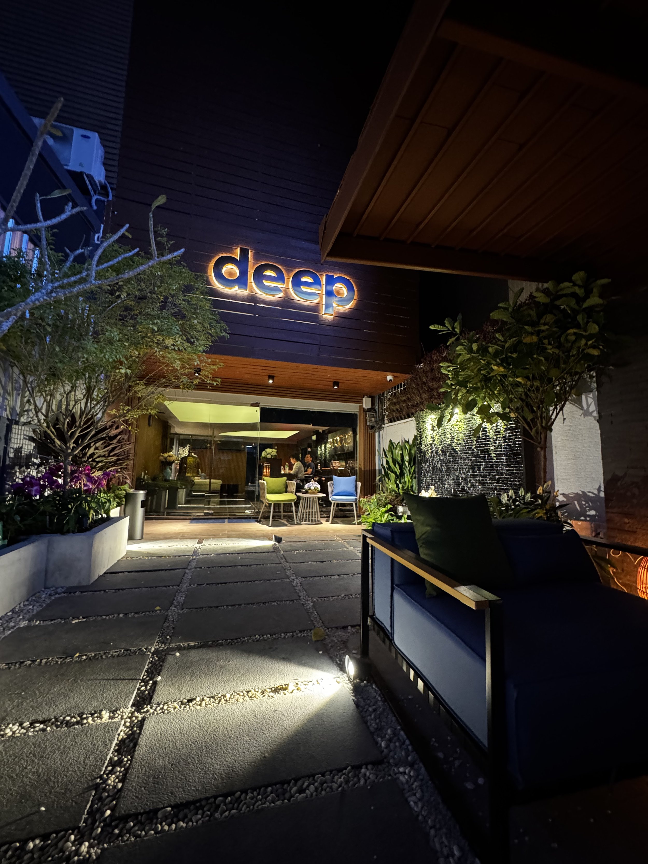 Night view of the exterior of a building with a sign that says 'deep' illuminated on the dark wooden wall. There are chairs and tables outside with plants and flowers, creating a cozy outdoor seating area.